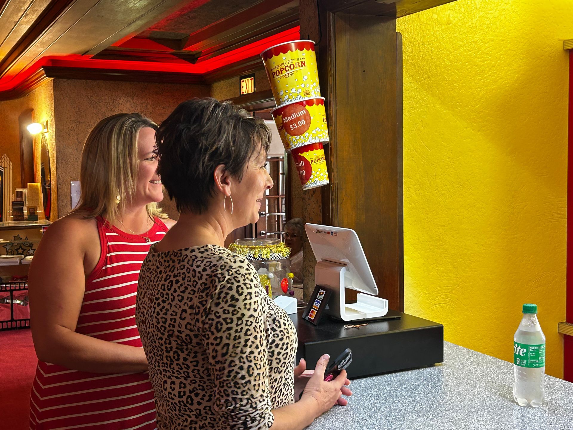 Two people stand at a service counter in a theater with popcorn tubs hanging in the background and a water bottle on site.