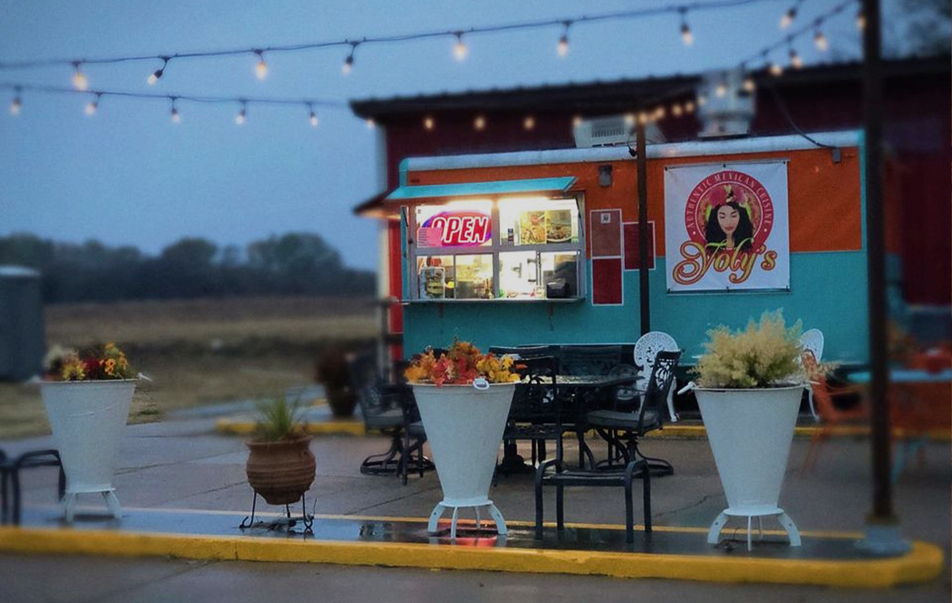 A teal and orange food truck, branded with a circular logo, sits outdoors at dusk under a string of glowing lights.