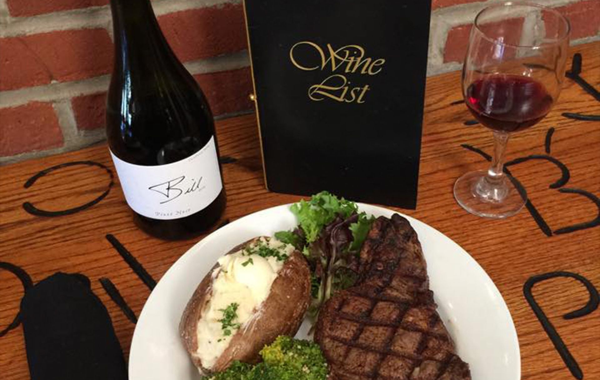 A grilled steak and baked potato on a white plate, served with a glass of red wine and a bottle on a wooden table.
