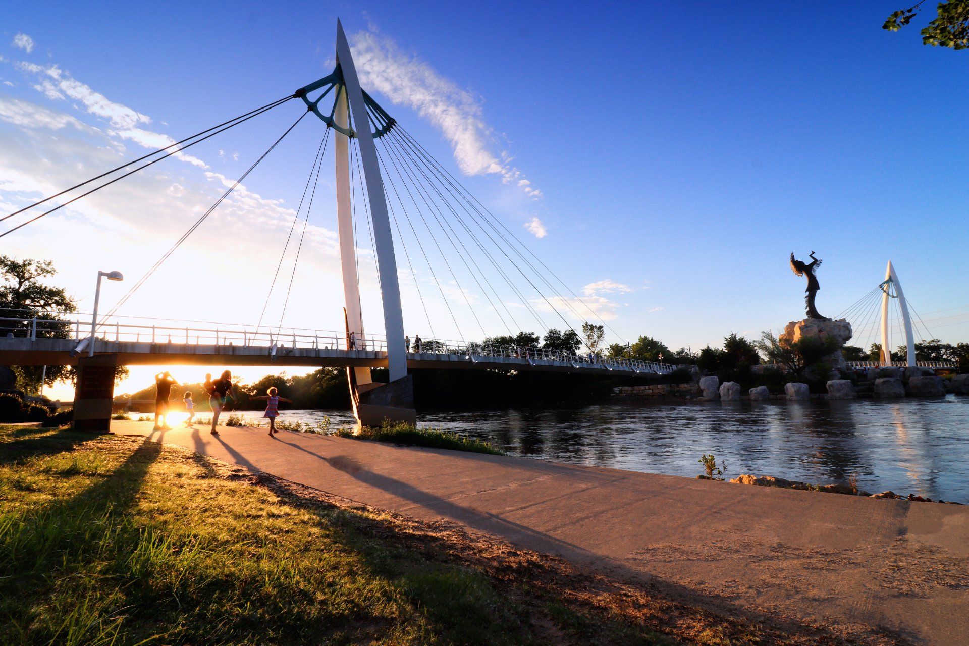 A sunlit pedestrian bridge and a tall statue of a person stand over a river, with silhouettes of people on the bank.