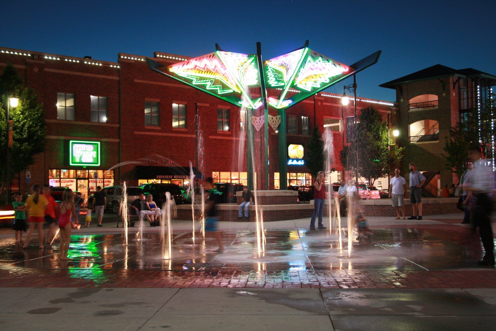 People enjoy a lit-up fountain in a public plaza at dusk, with brick buildings and illuminated signs in the background.