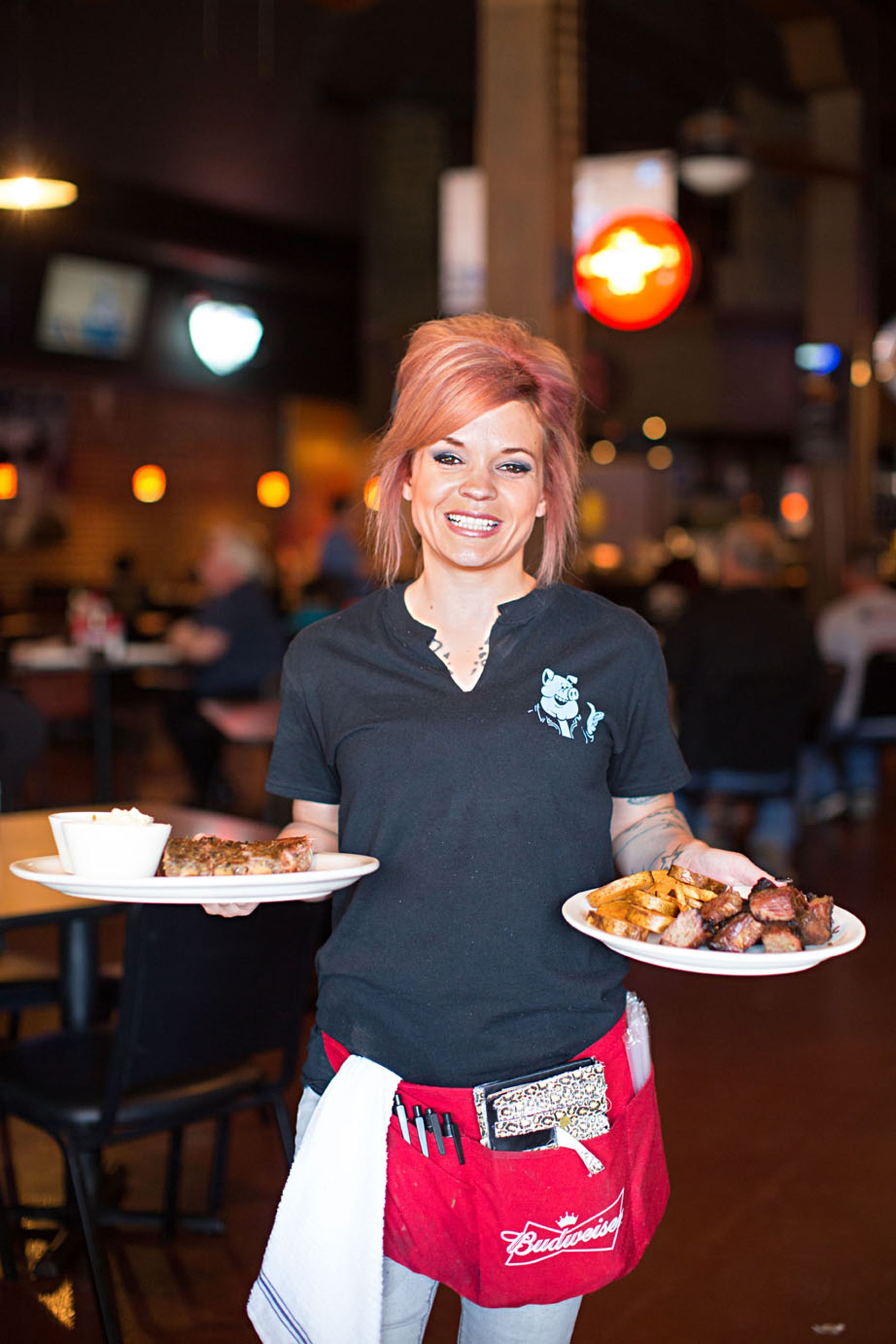 A smiling server in a black shirt and red apron holds two plates of food in a restaurant dining area.