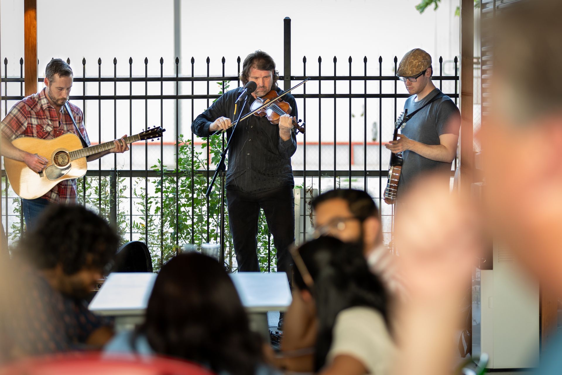 Three musicians play an acoustic set on a patio, viewed from behind an audience seated at tables.