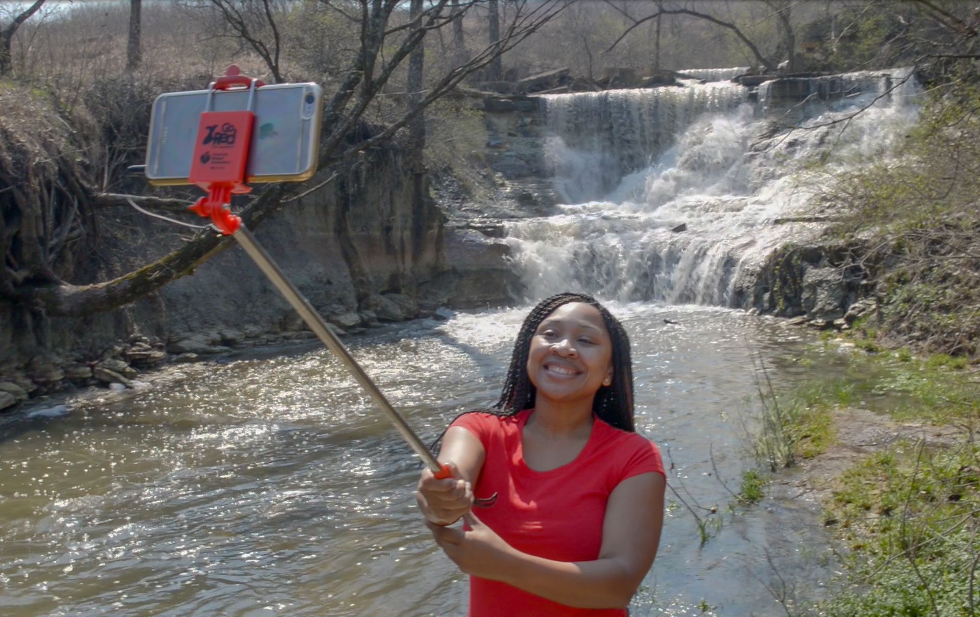 A person in a red shirt smiles while taking a selfie with a pole in front of a waterfall in a wooded area.