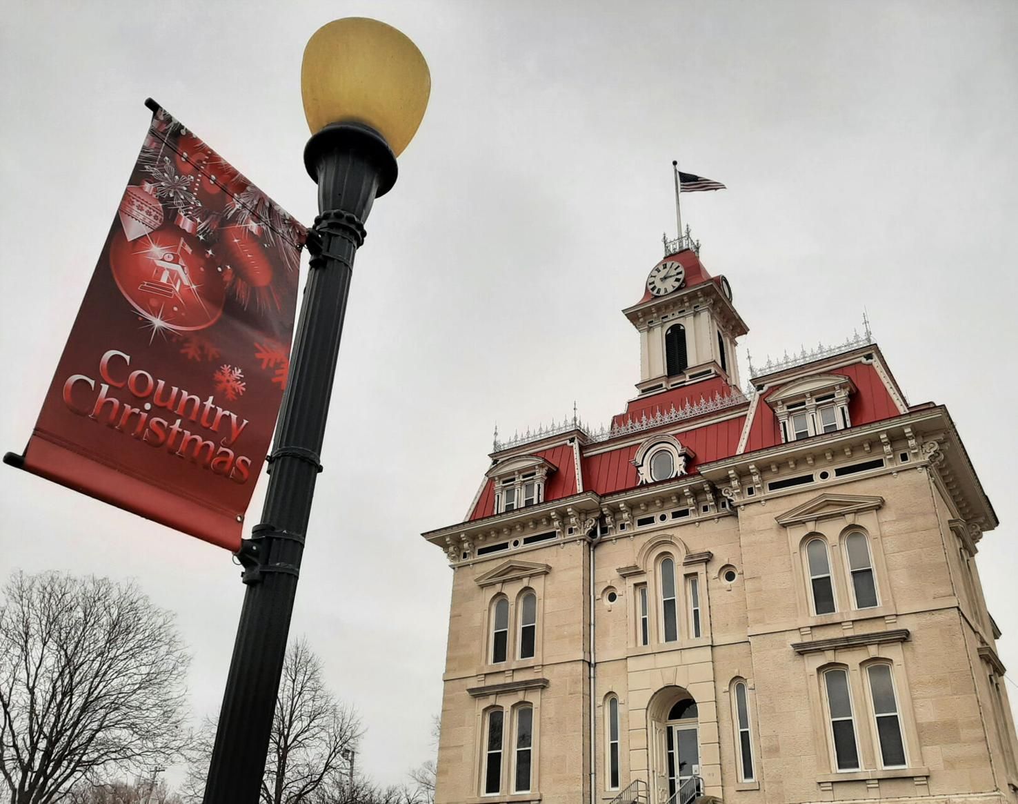 A red Country Christmas banner hangs on a lamp post in front of a historic stone courthouse with a red roof and clock.