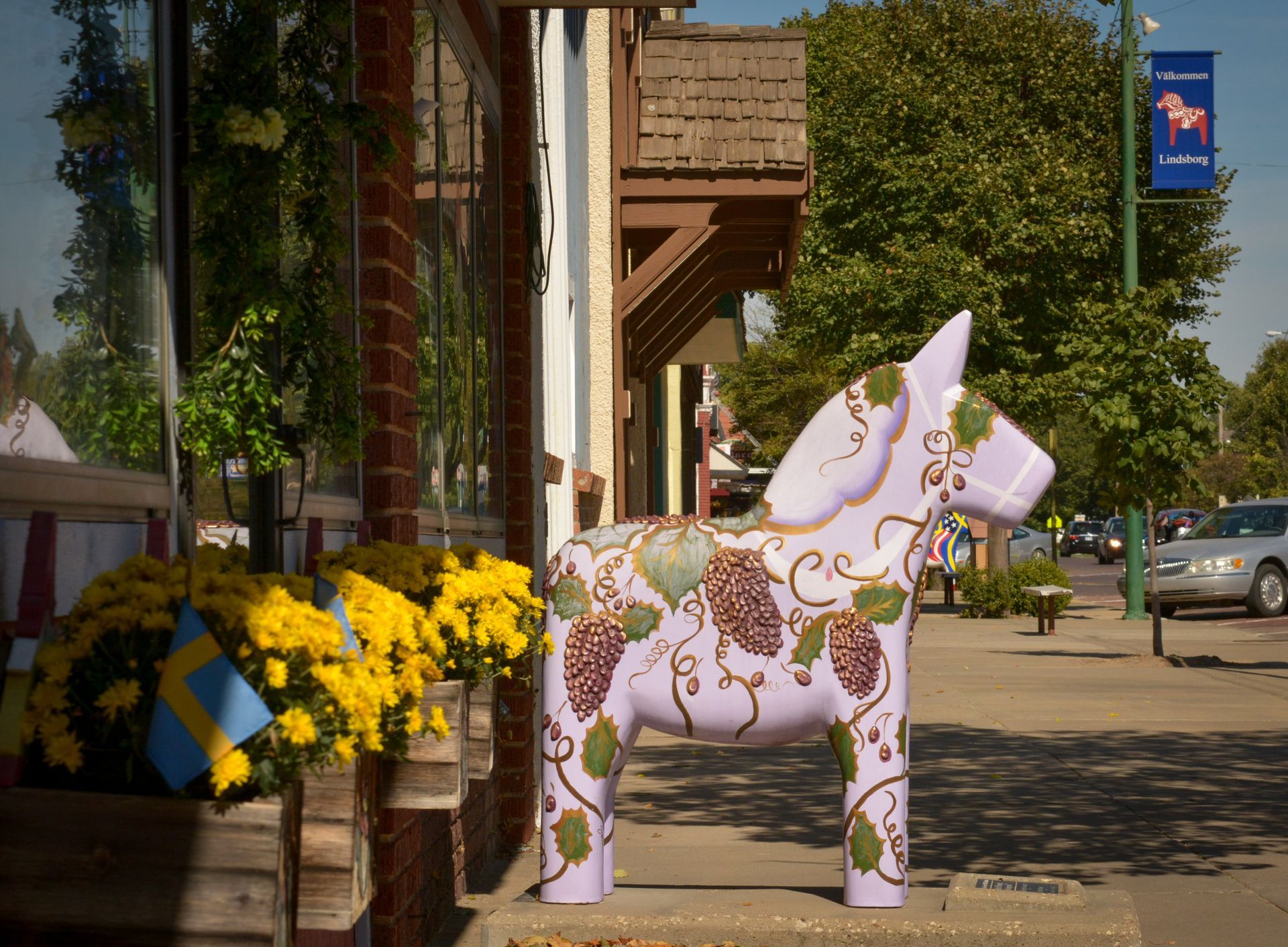 A light purple painted Dala horse statue sits outside a shop with yellow flowers and a flag in Lindsborg, Kansas.