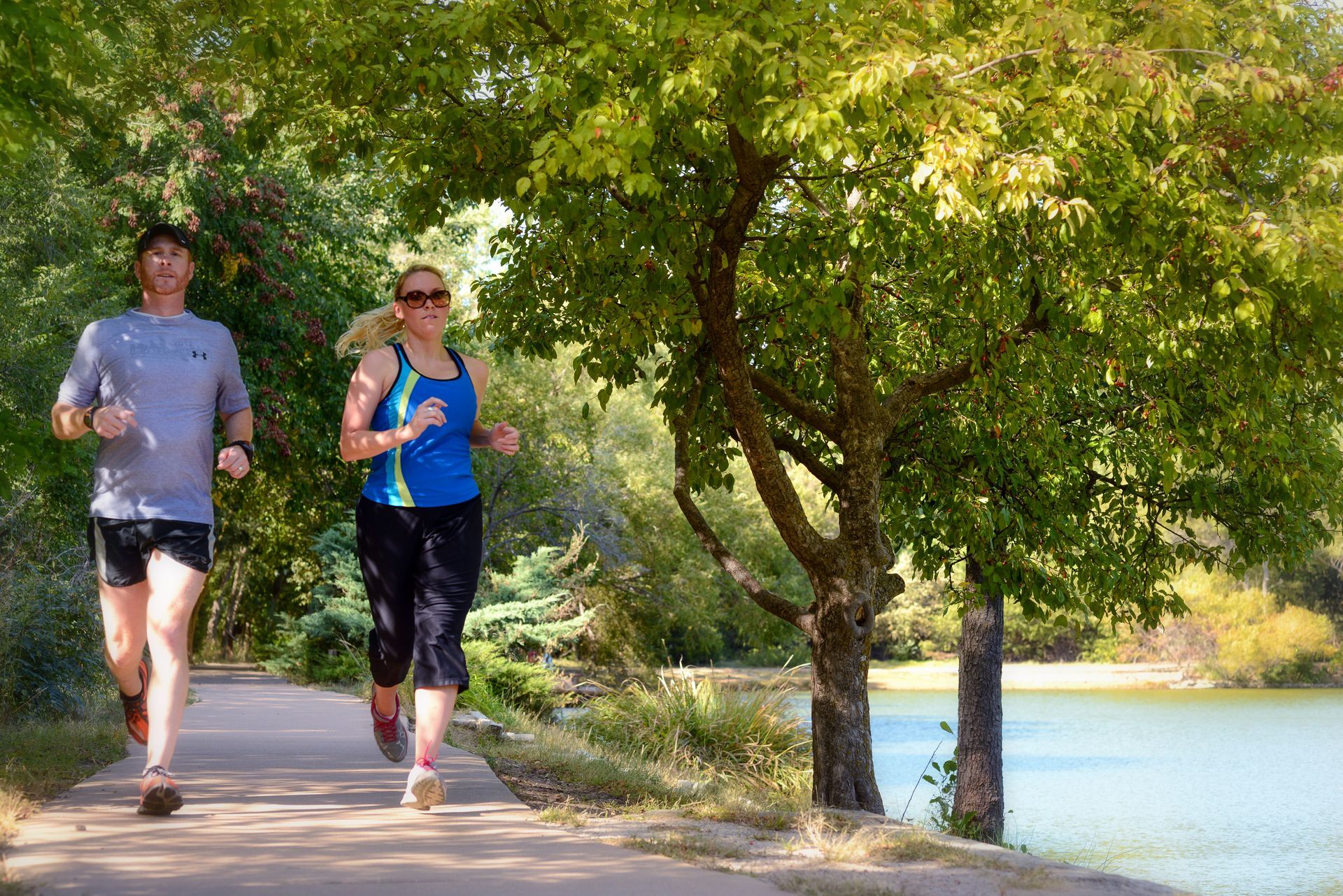 Two runners jog along a paved trail beside a tree-lined lake on a sunny day.