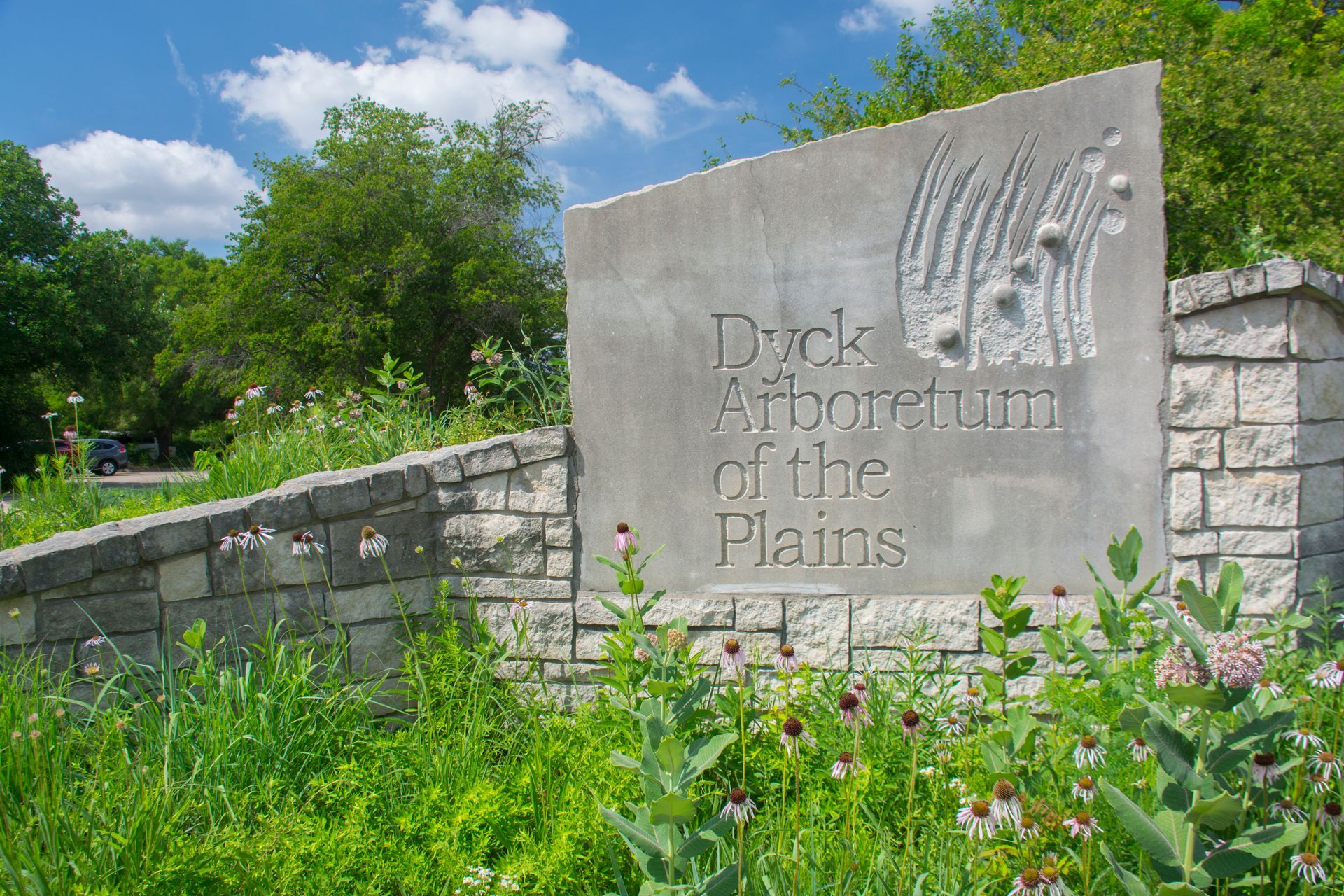 Stone sign for Dyck Arboretum of the Plains surrounded by lush green prairie plants under a sunny blue sky.