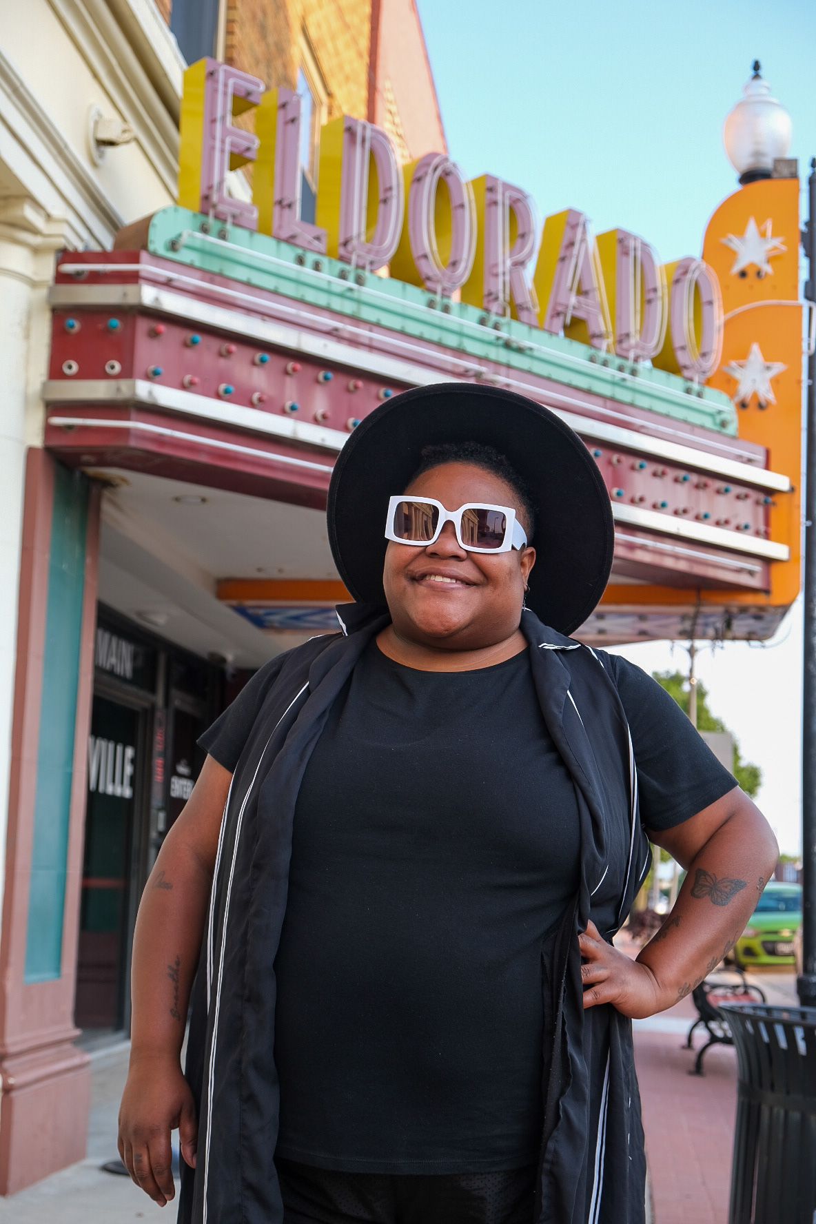 A smiling person in a black wide-brimmed hat and white-framed sunglasses stands before the Eldorado theater marquee.