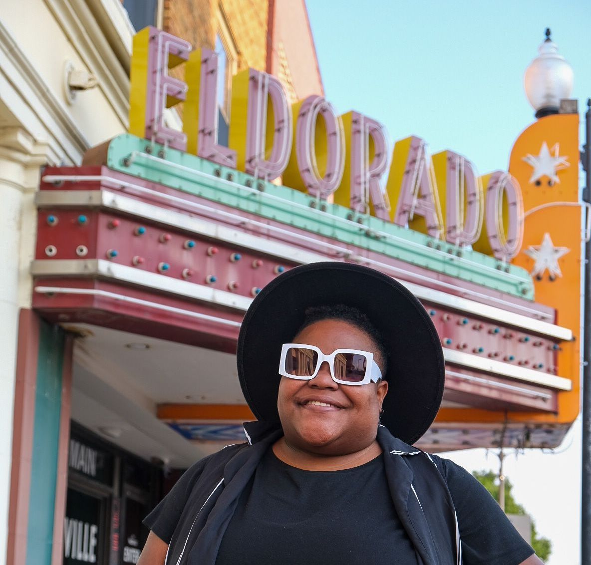 A smiling person wearing a black wide-brimmed hat and white-framed sunglasses stands before the Eldorado theater sign.
