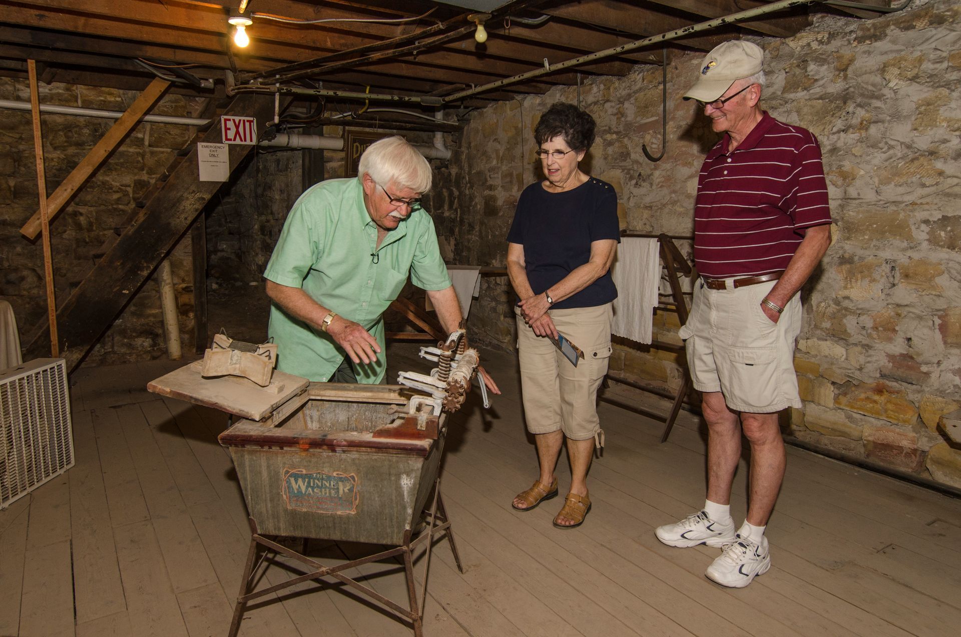 A person demonstrates a vintage metal washbasin to two onlookers in a stone-walled cellar.