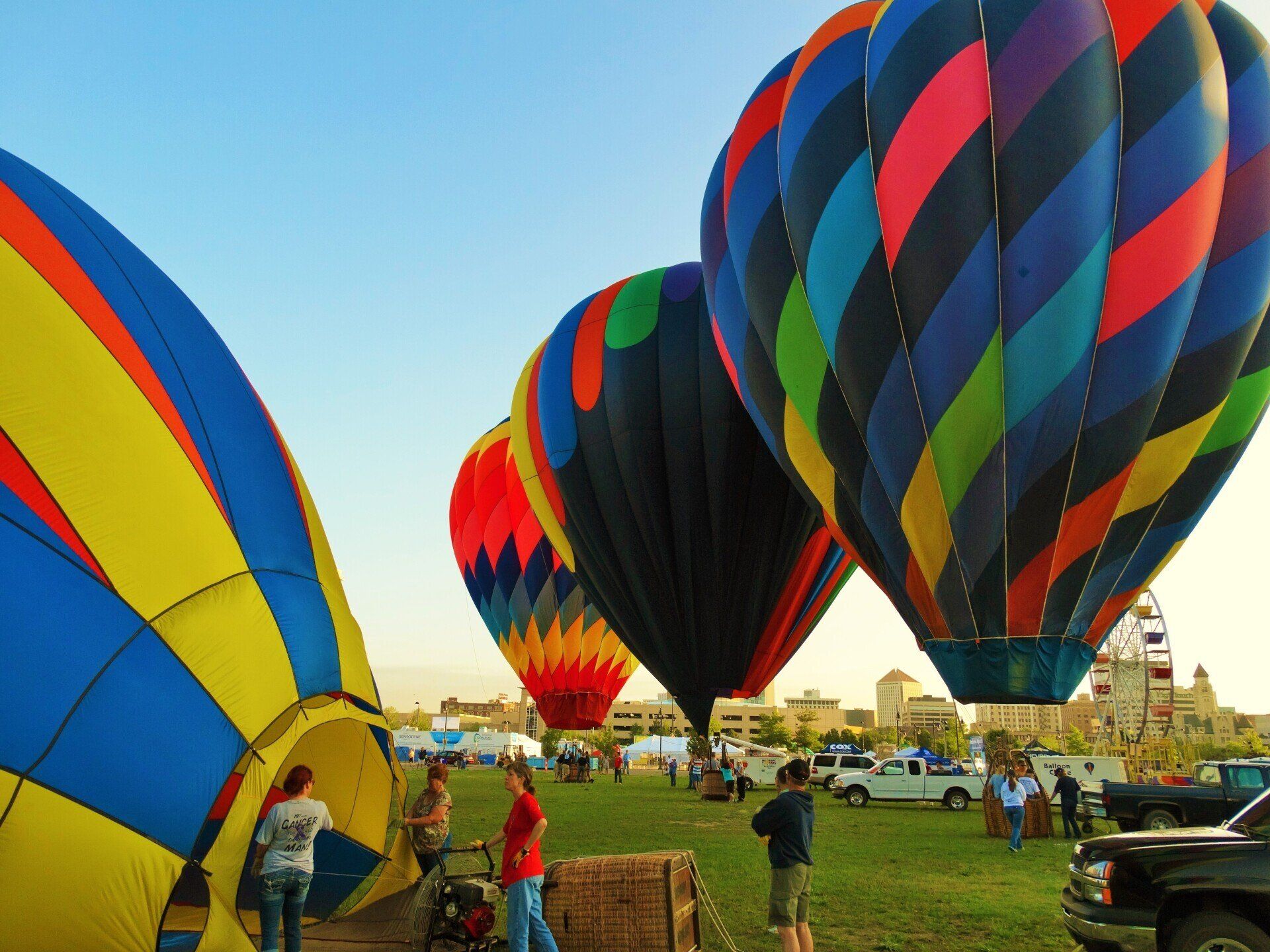 Multiple colorful hot air balloons sit on a grassy field at a festival, with people preparing them for flight.