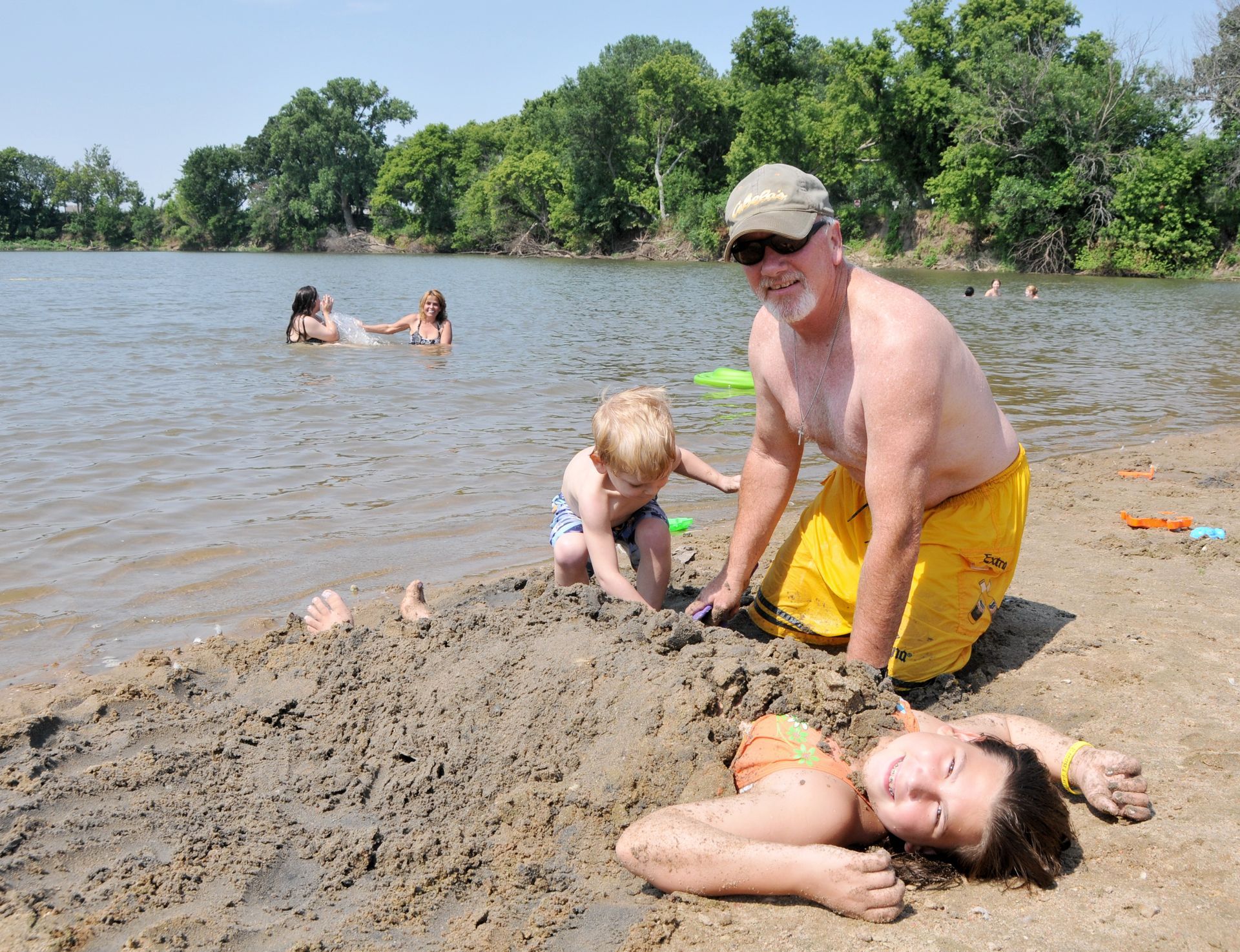 A person in yellow shorts and a child play in the sand on a beach, partially burying another person, near a lake.