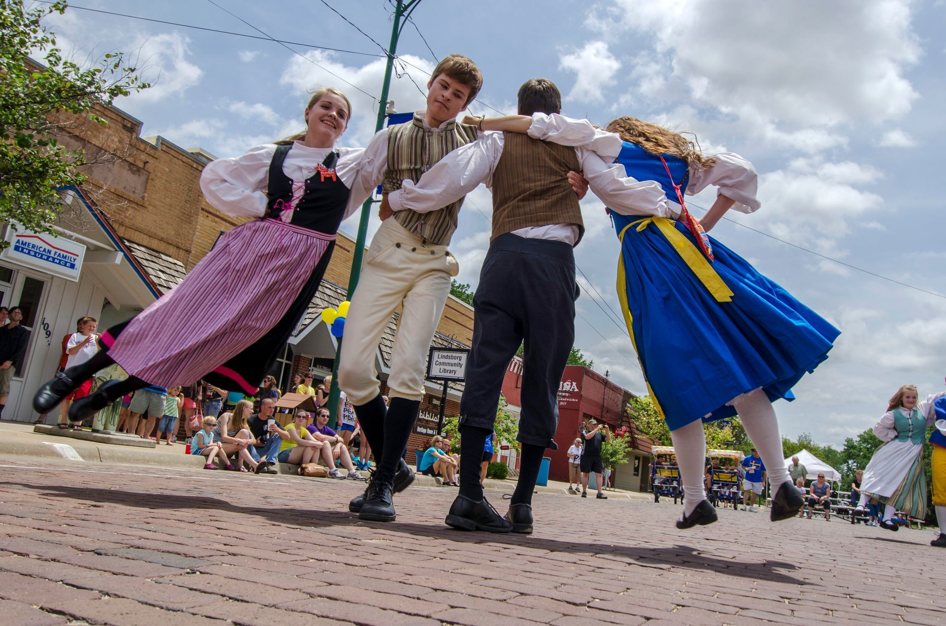Four people in traditional folk clothing dance in a circle on a brick street during an outdoor public event.