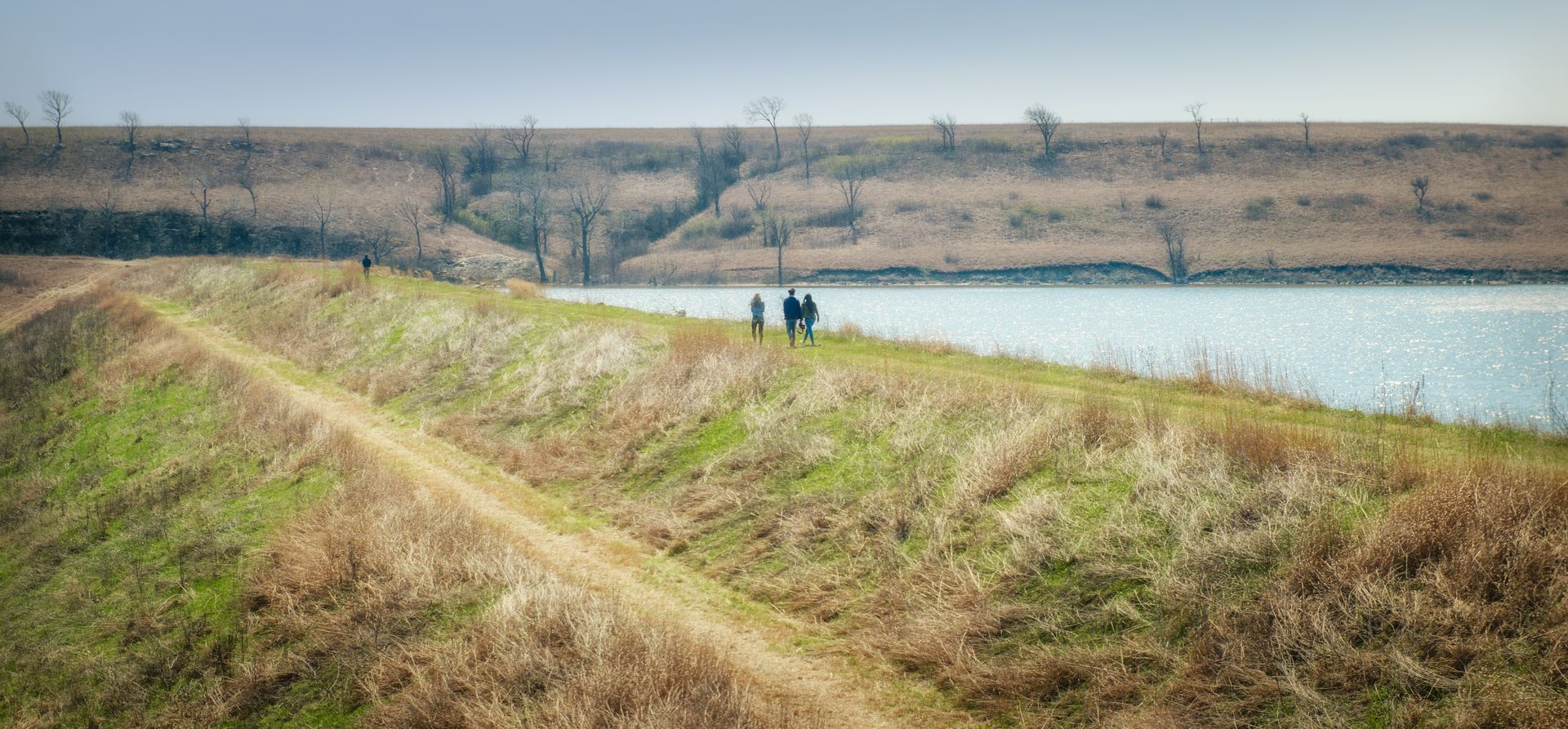 Three people walk along a dirt path on a grassy levee beside a body of water under a pale sky.