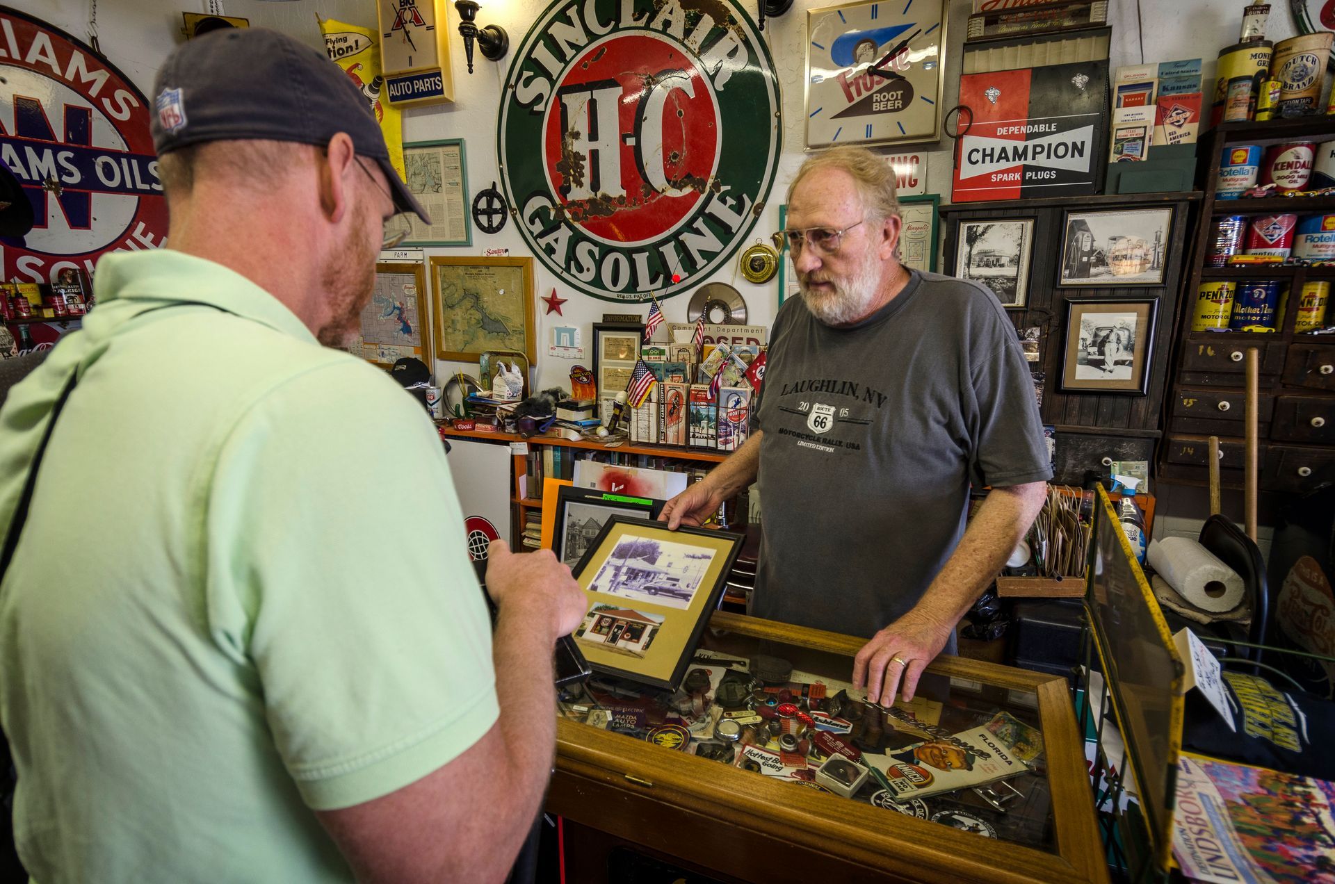 A man in a light green shirt looks at a display case while a bearded man in a gray shirt assists him in a vintage shop.