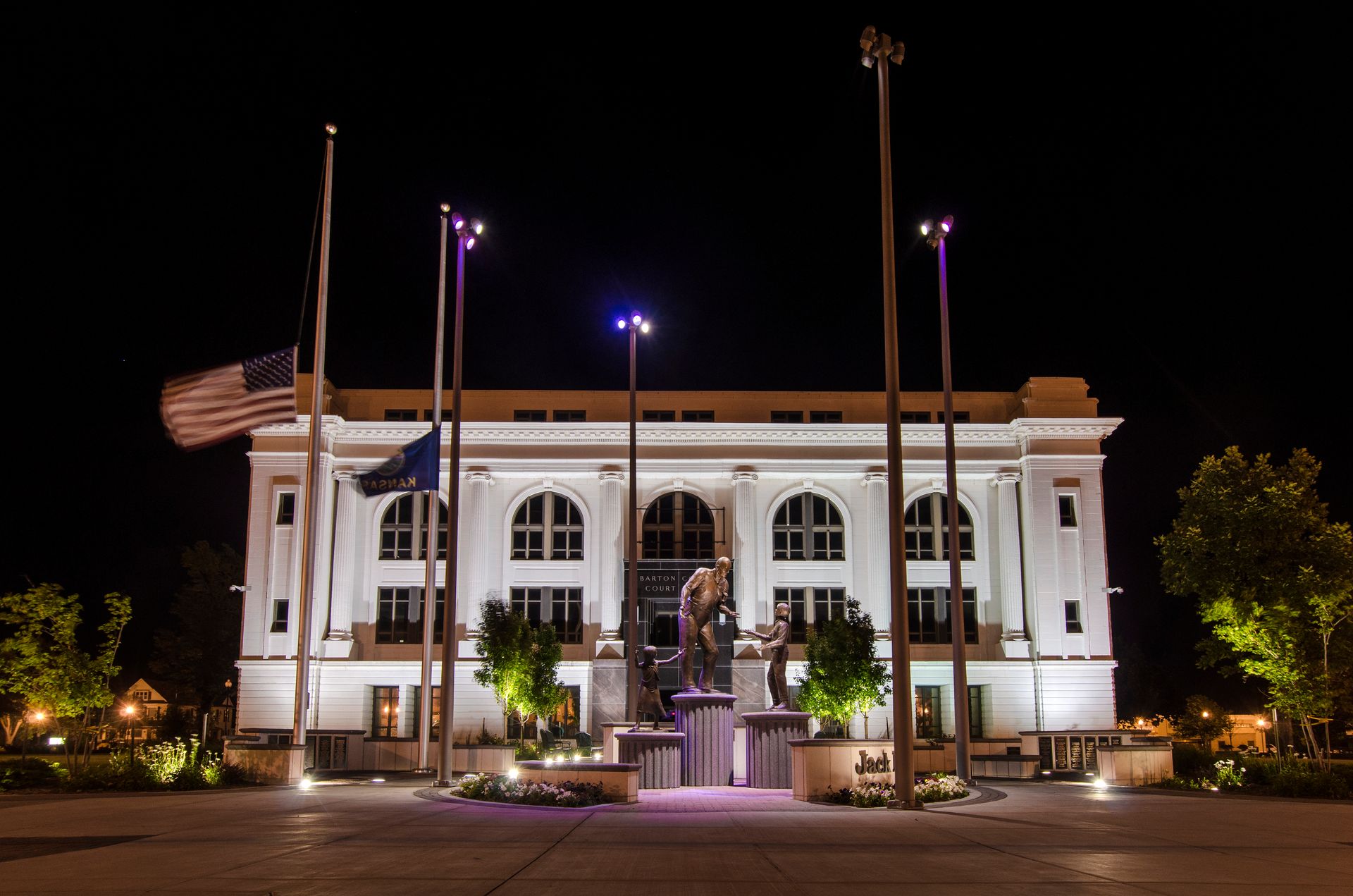 A white building with arched windows and a central monument is illuminated at night, with two flags flying at half-mast.