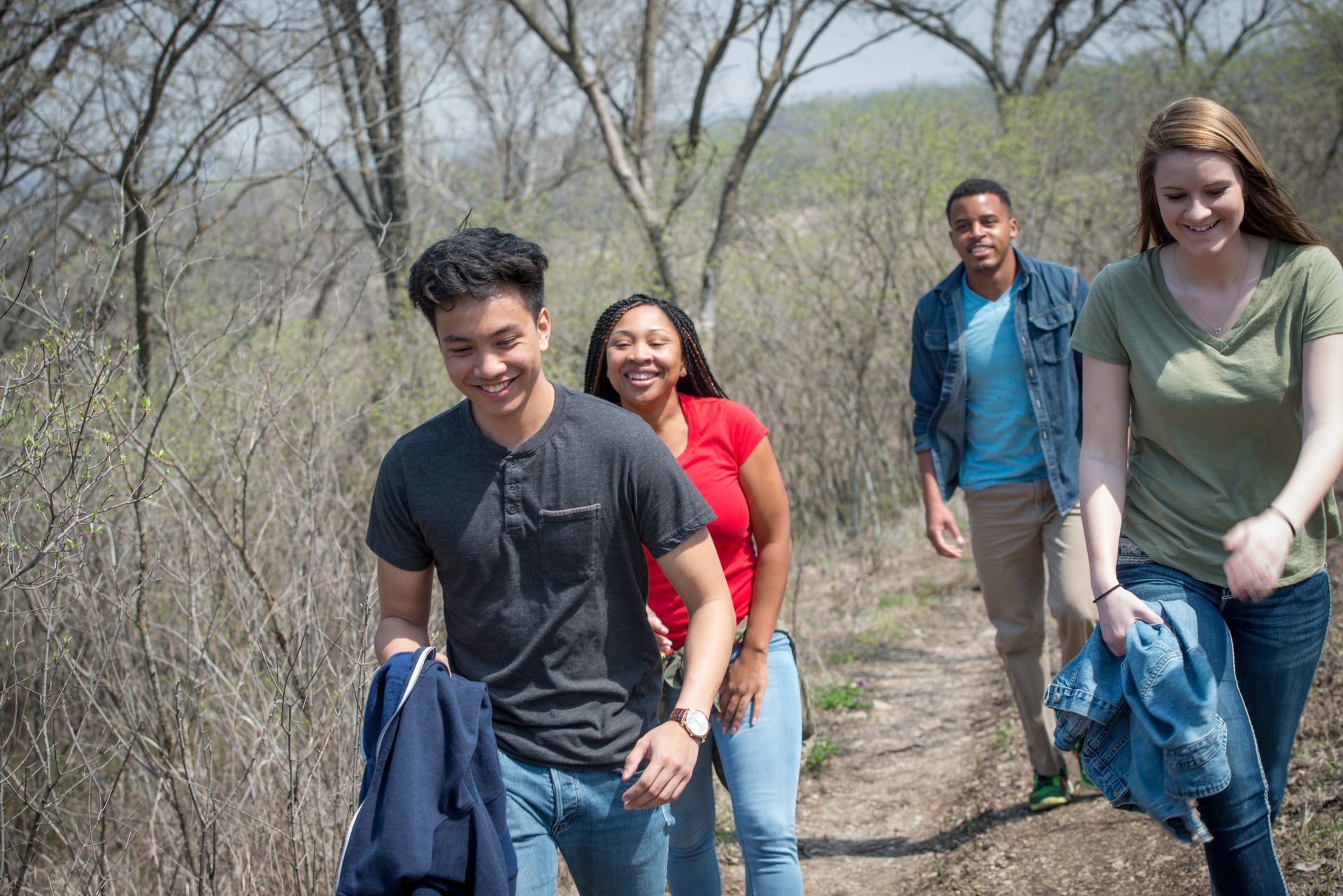 Four people smiling while hiking together on a narrow dirt trail through a lightly wooded forest.