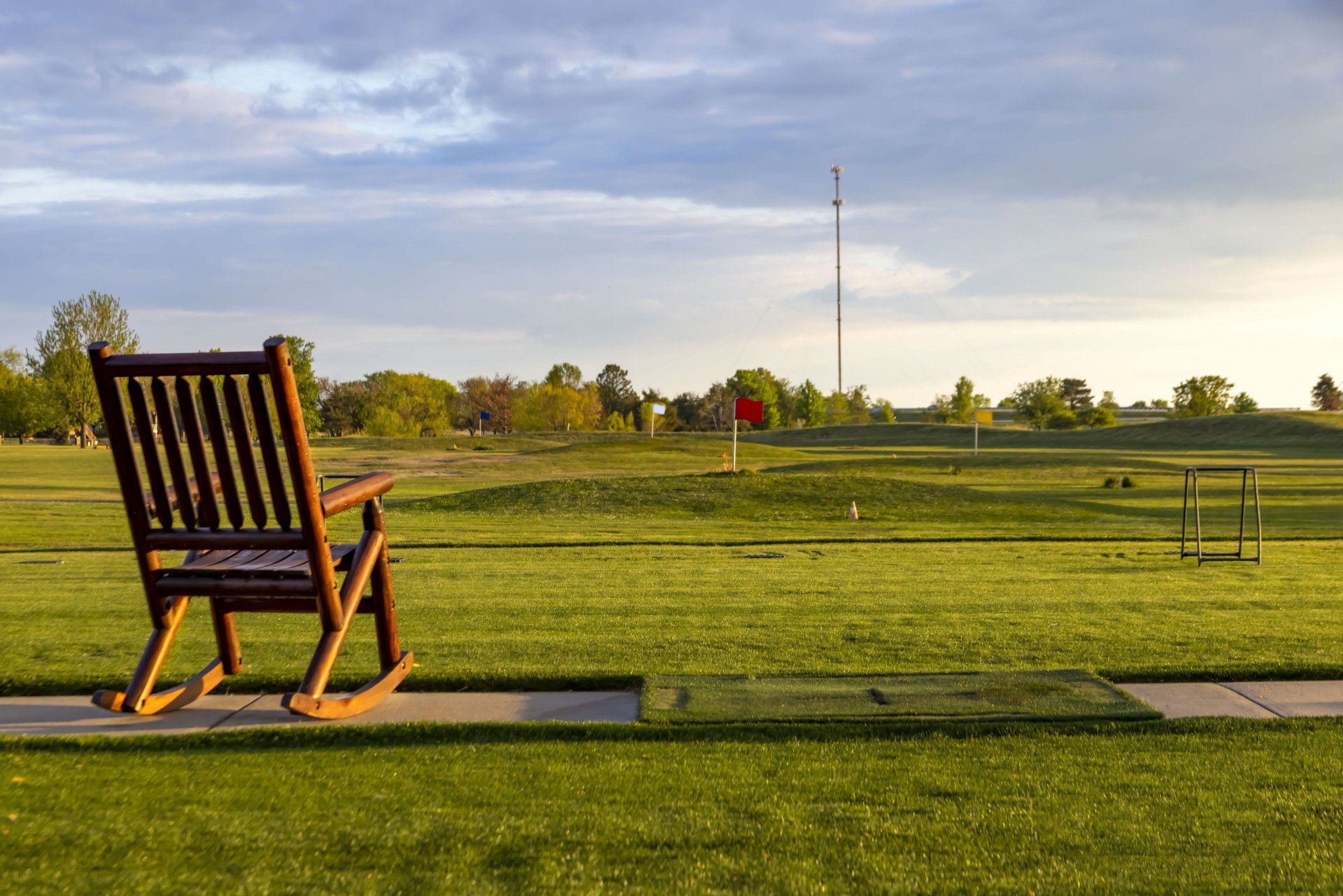 A wooden rocking chair sits on a concrete path overlooking a grassy golf driving range at sunset.