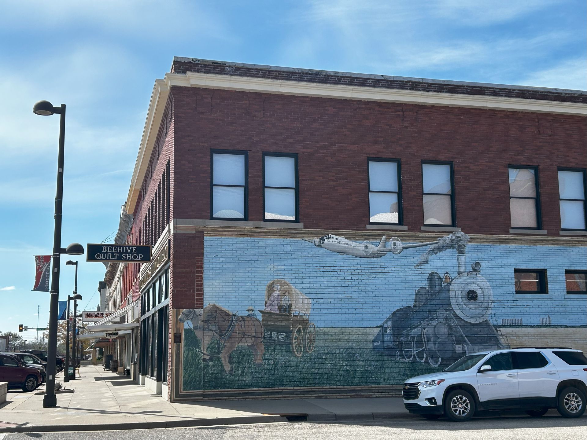 A brick building with a large mural of a train and covered wagon, with a white SUV parked in front on a sunny day.