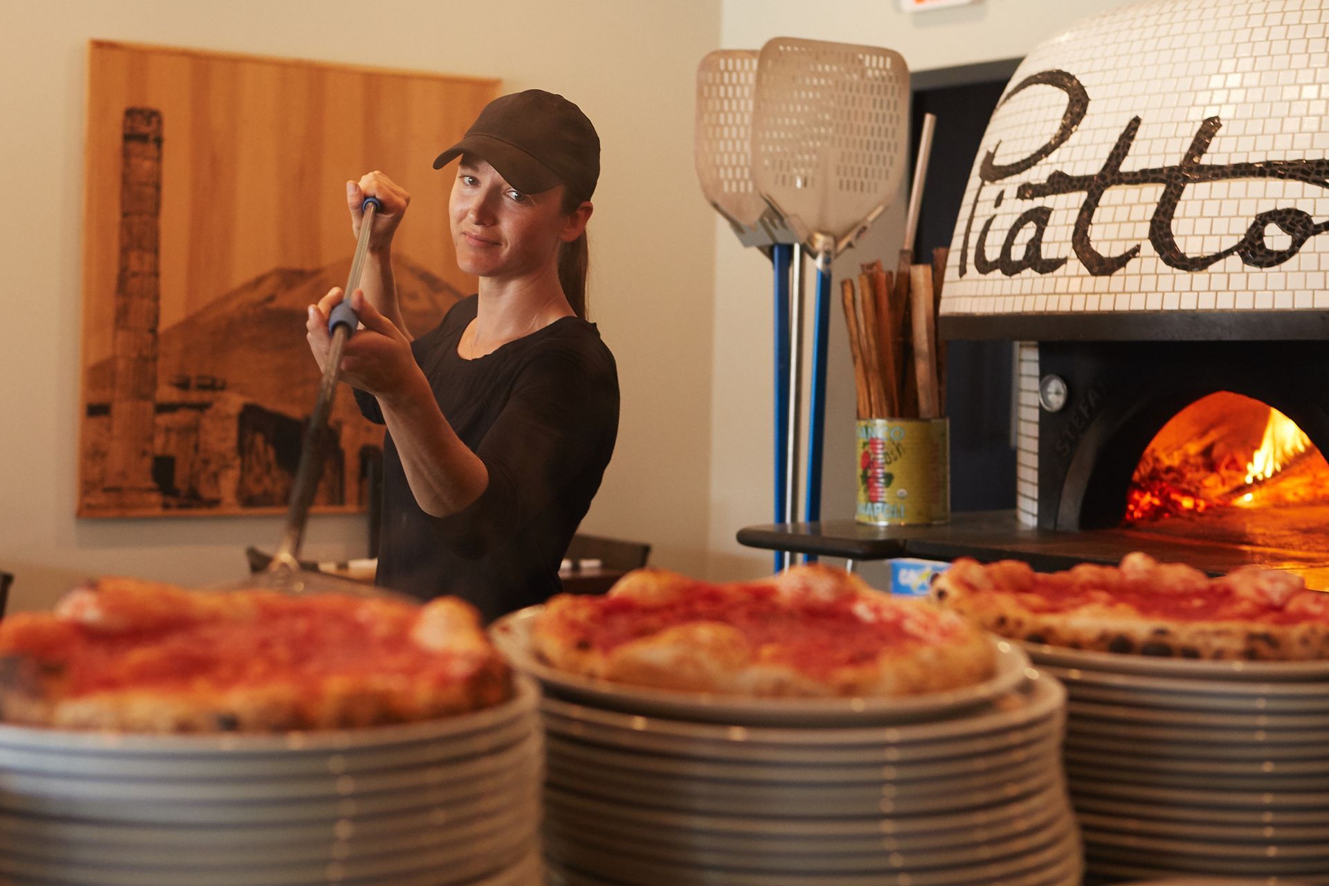A cook stands in a kitchen using a long peel near a fired-up pizza oven, with stacks of finished pizzas in the foreground.