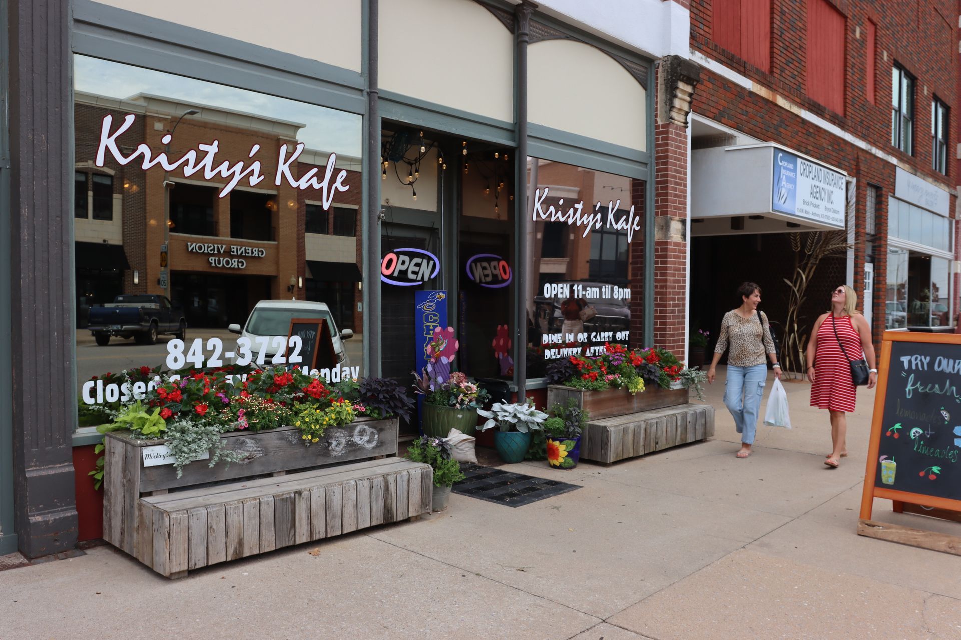 Two people walk past the exterior of Kristy's Kafe on a sidewalk with wooden planter boxes and an outdoor sign.