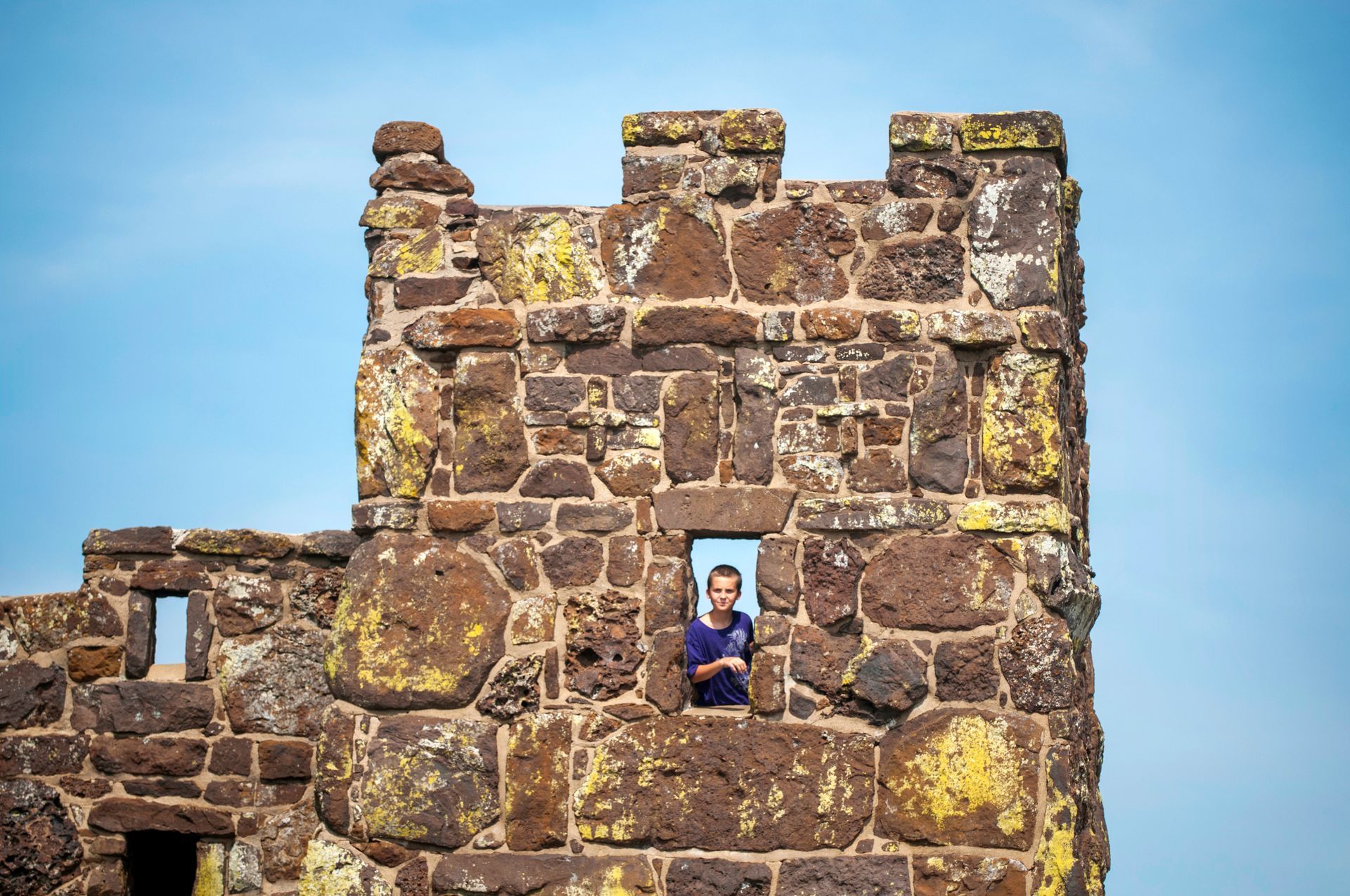A person in a blue shirt looks out from a window in a stone castle tower against a blue sky outside of Lindsborg, Kansas.
