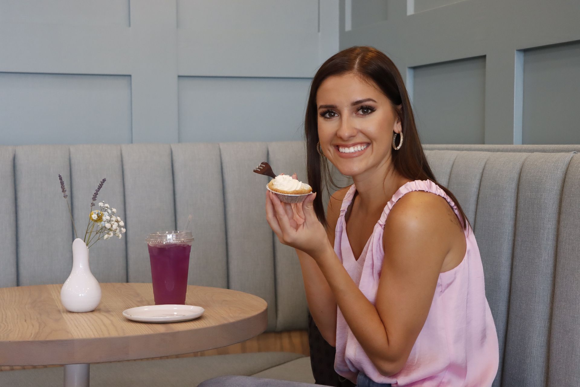 A smiling person in a light pink top holds a small pastry while seated in a booth with a purple drink and flower vase.