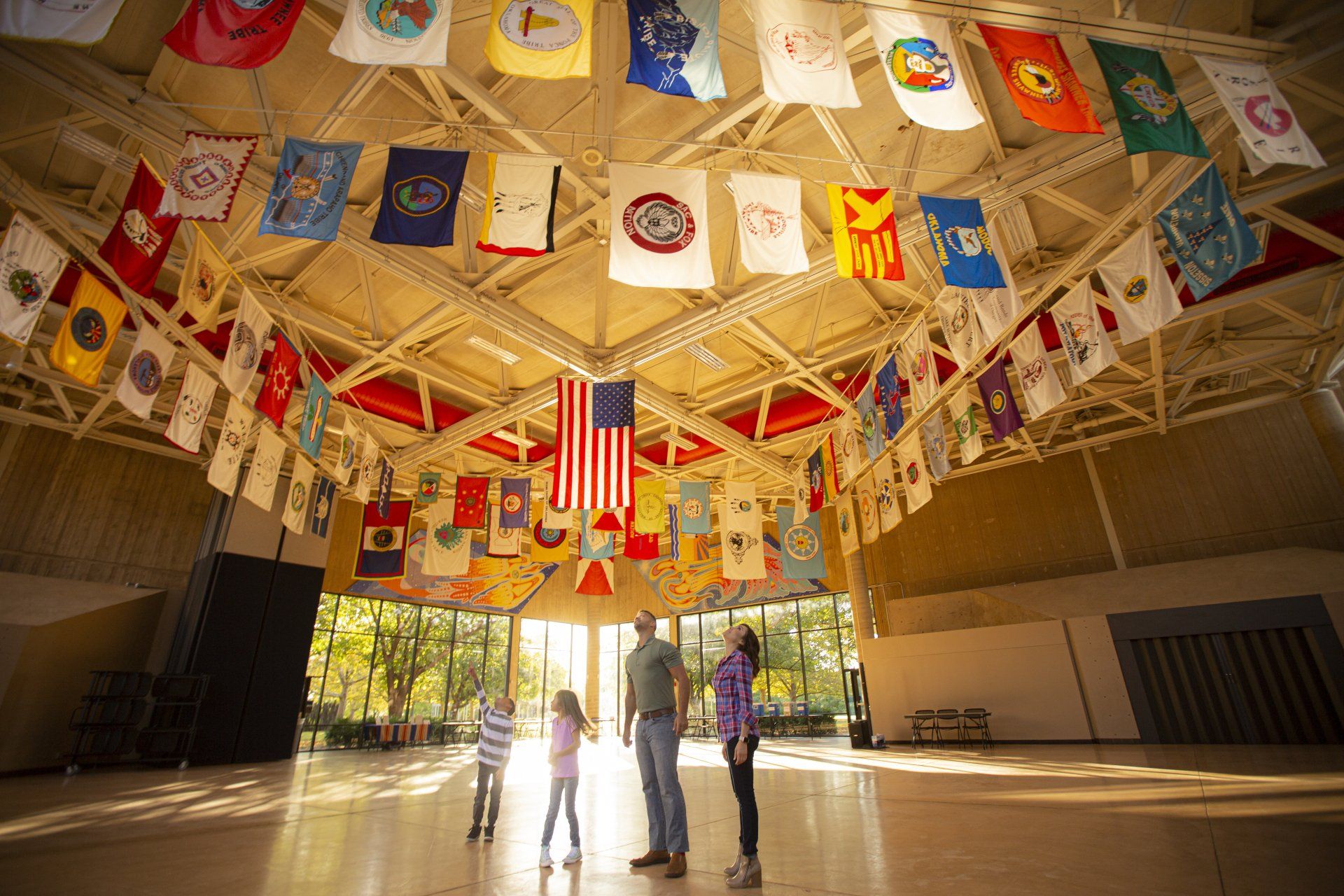 People stand in a hall with a high ceiling decorated with numerous flags, featuring a central American flag.