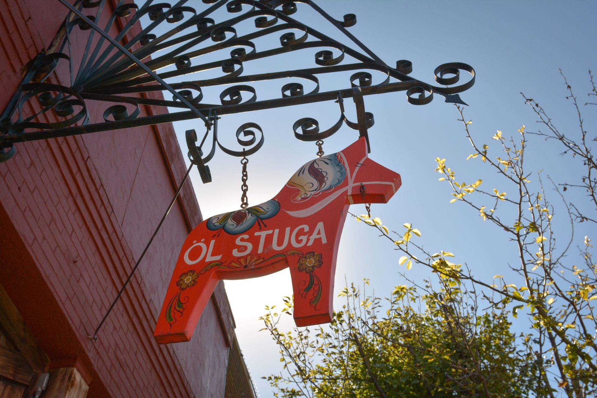 A red, hand-painted wooden Dala horse sign hangs from an ornate metal bracket against a clear blue sky.