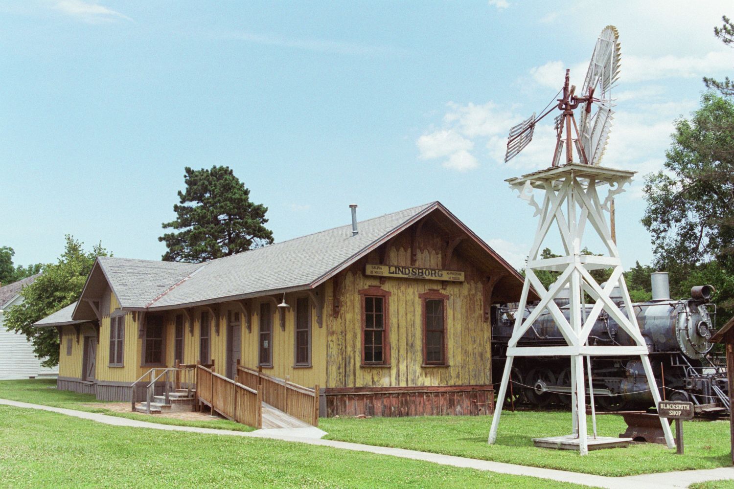 A yellow historic train depot with a wooden ramp, next to a large white windmill and a vintage steam locomotive.