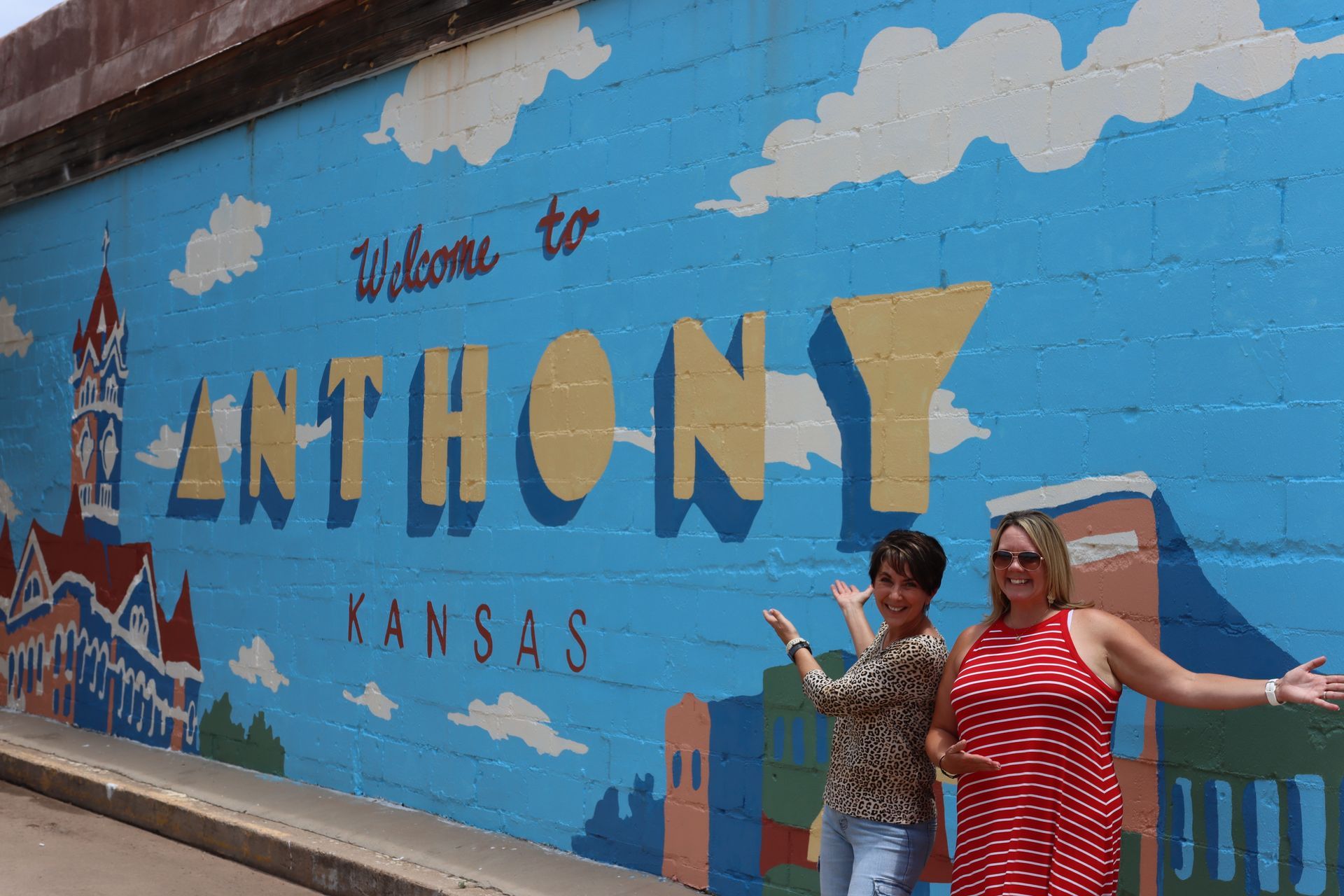 Two people stand smiling and gesturing in front of a blue mural that reads, 