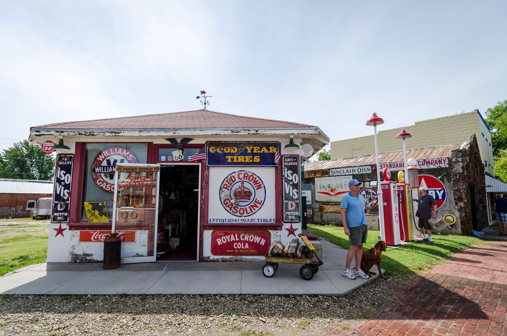 A vintage-style roadside shop with retro signage stands outdoors, with a person and a small dog standing nearby.