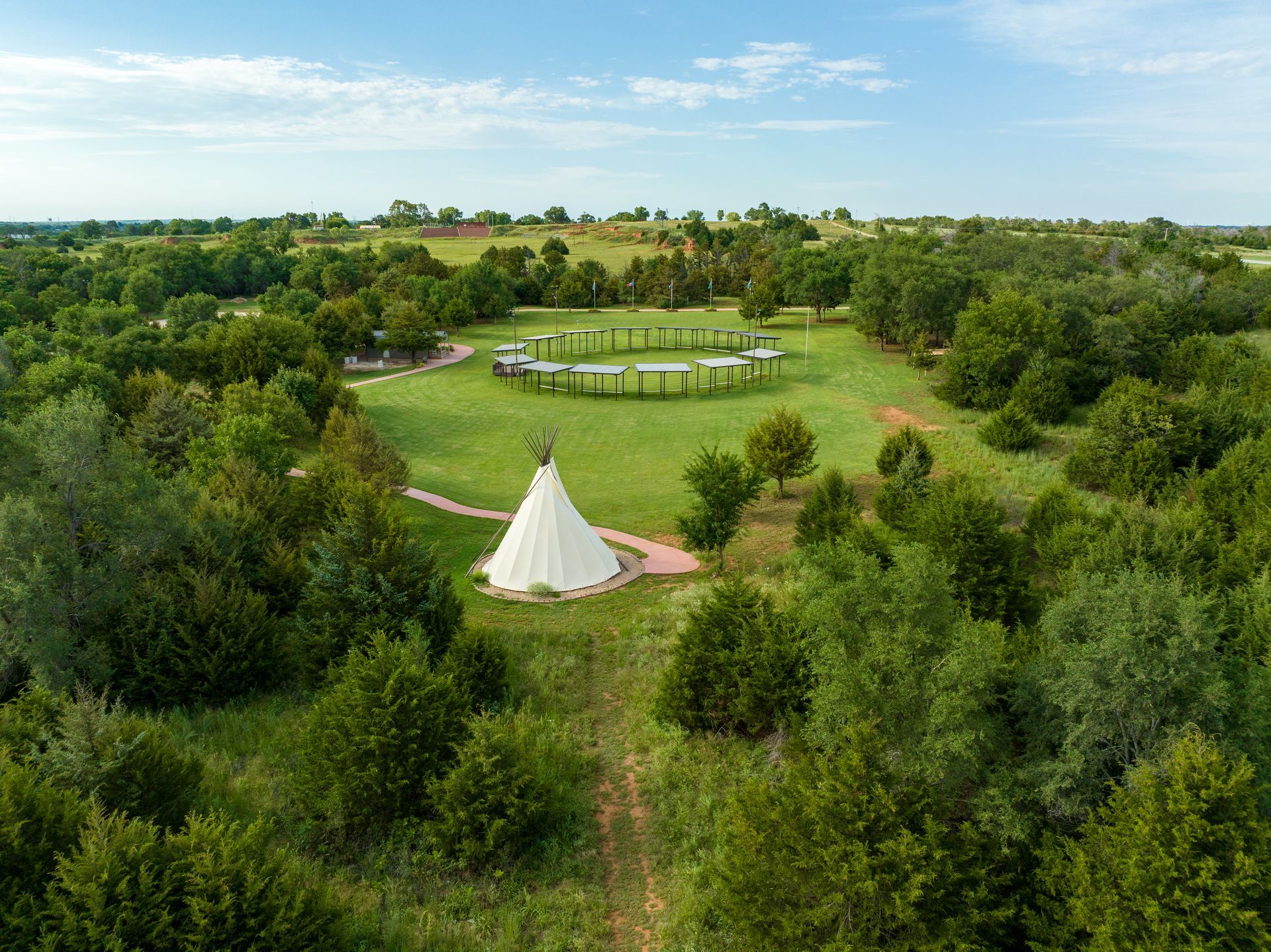 A white tipi sits in a lush green field, with a circular wooden structure and dense forest in the background.