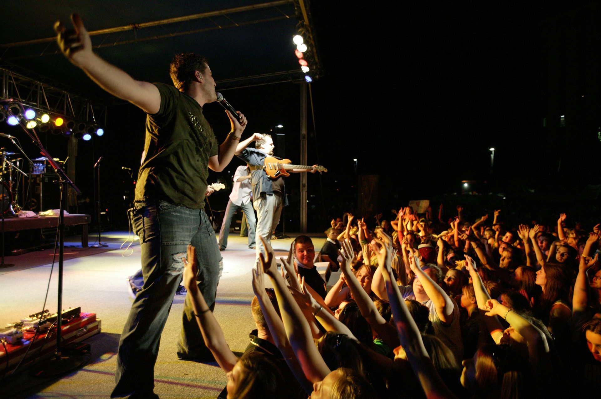 A singer performs on a stage at night with arms outstretched toward an enthusiastic crowd reaching back with hands raised.