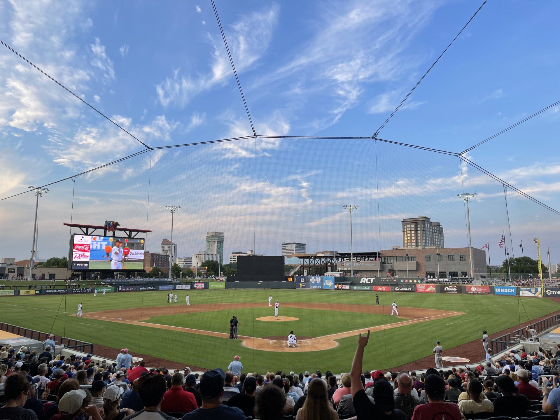 A baseball game in progress at a stadium under a blue sky, with fans in the foreground and city buildings in the distance.
