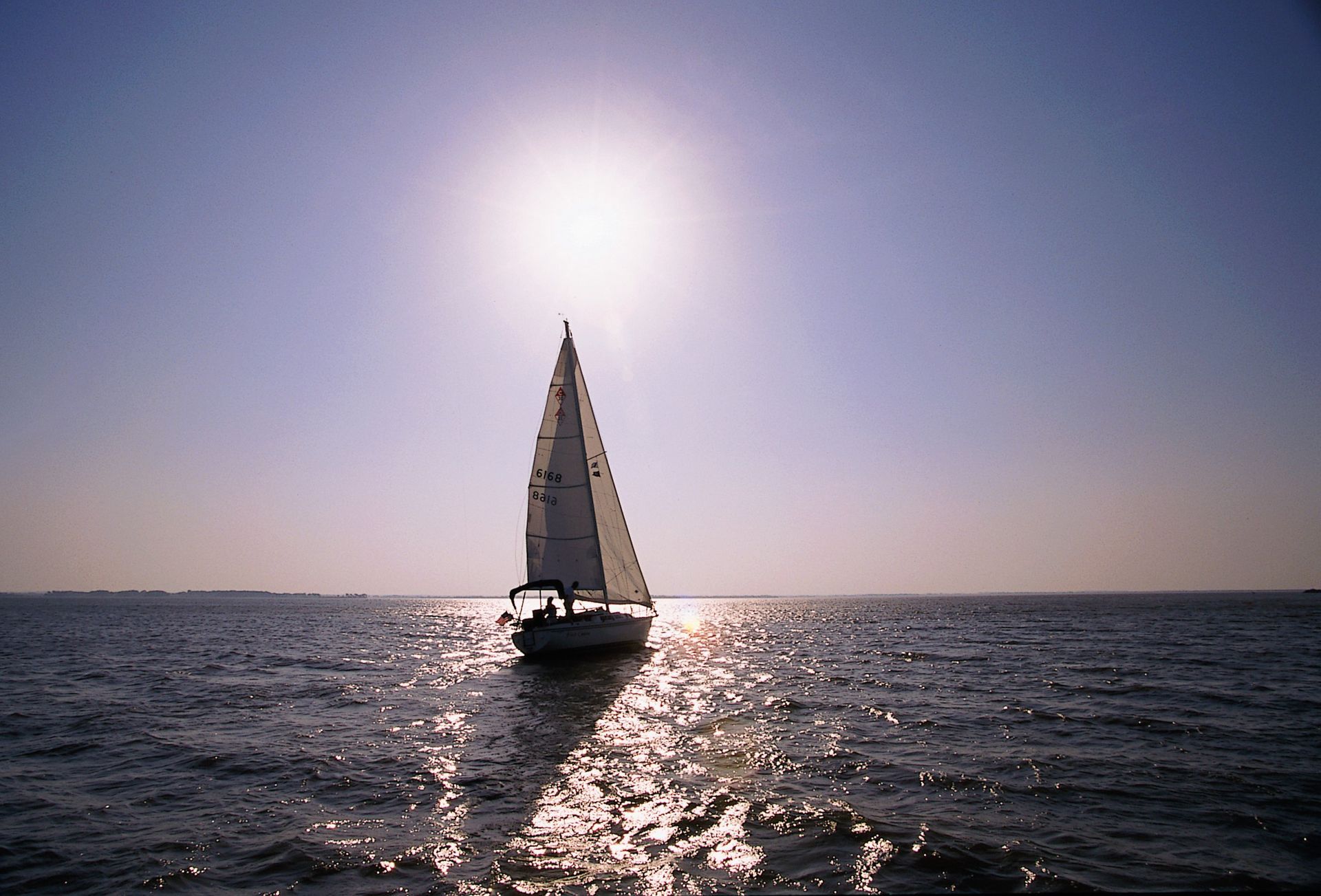 A sailboat is silhouetted against a bright sun over calm, shimmering water.
