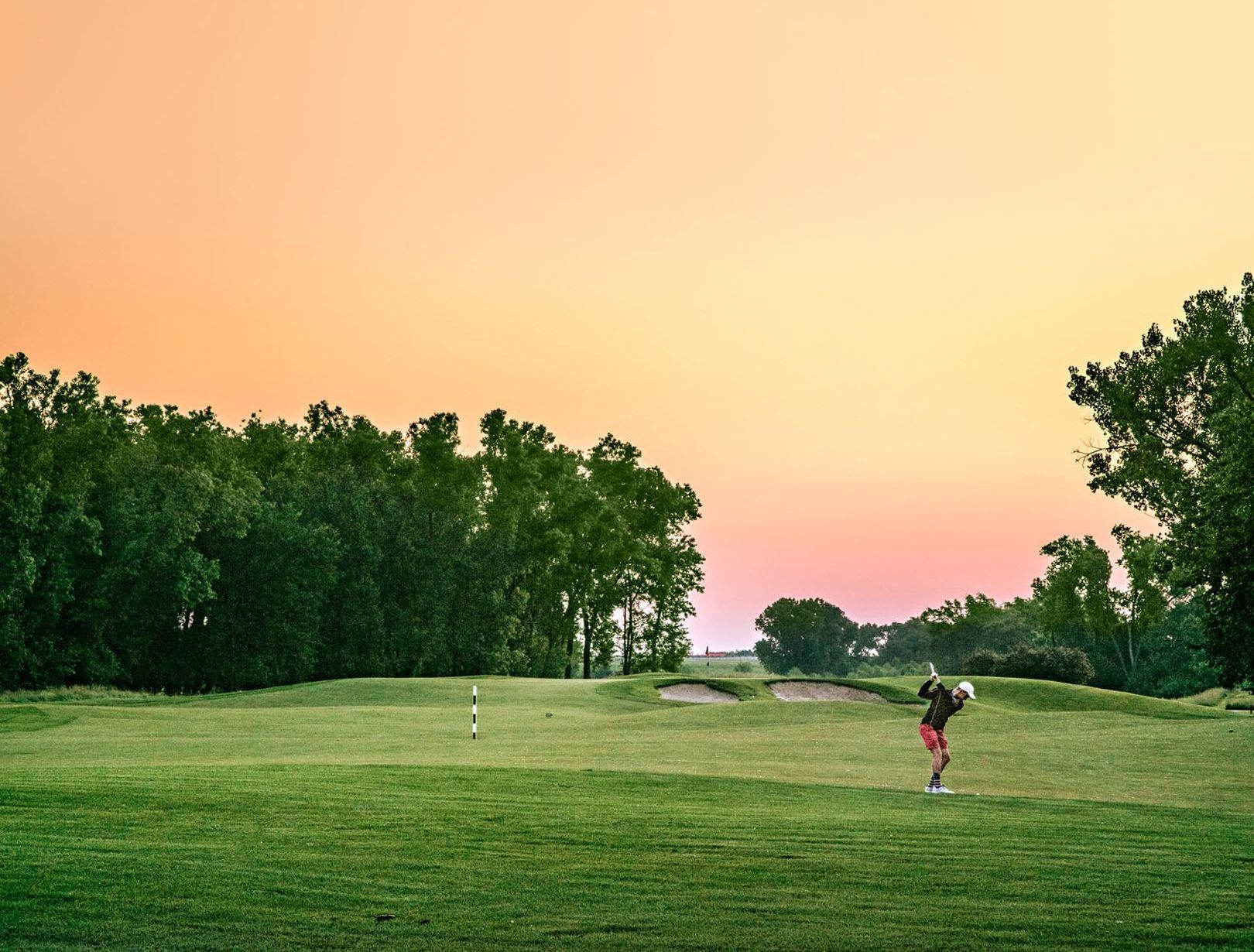A golfer takes a swing on a green course during a sunset with a gradient orange and pink sky in Newton, Kansas.