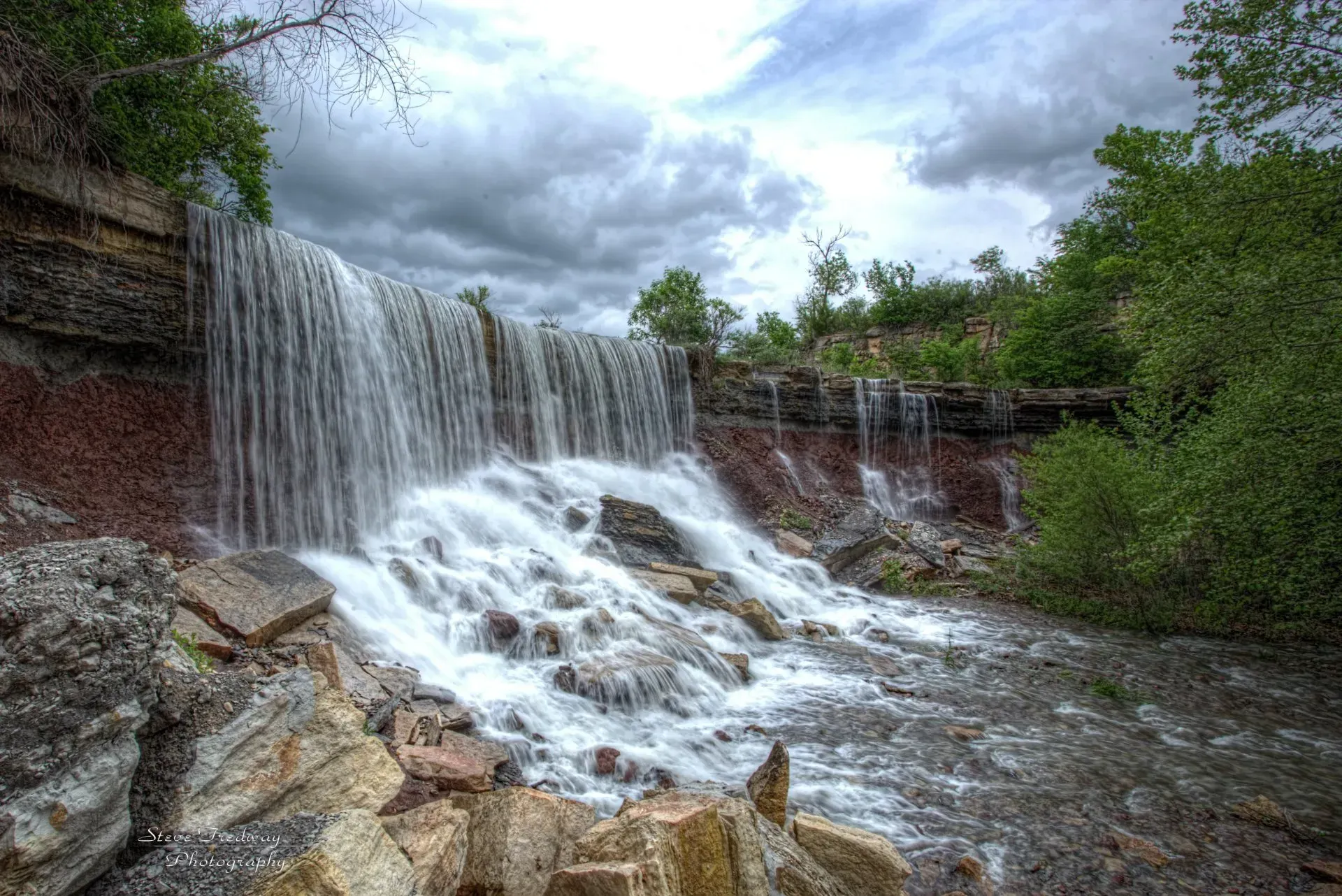 A wide, cascading waterfall flows over a layered, rock cliff into a rocky stream outside of Arkansas City, Kansas.