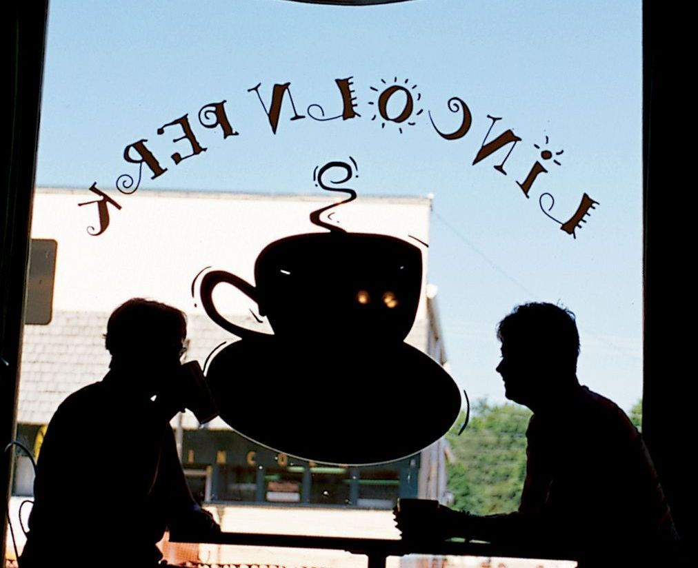 Silhouetted figures of two people sit at a table in a cafe, seen through a window featuring a Lincoln Perk logo.