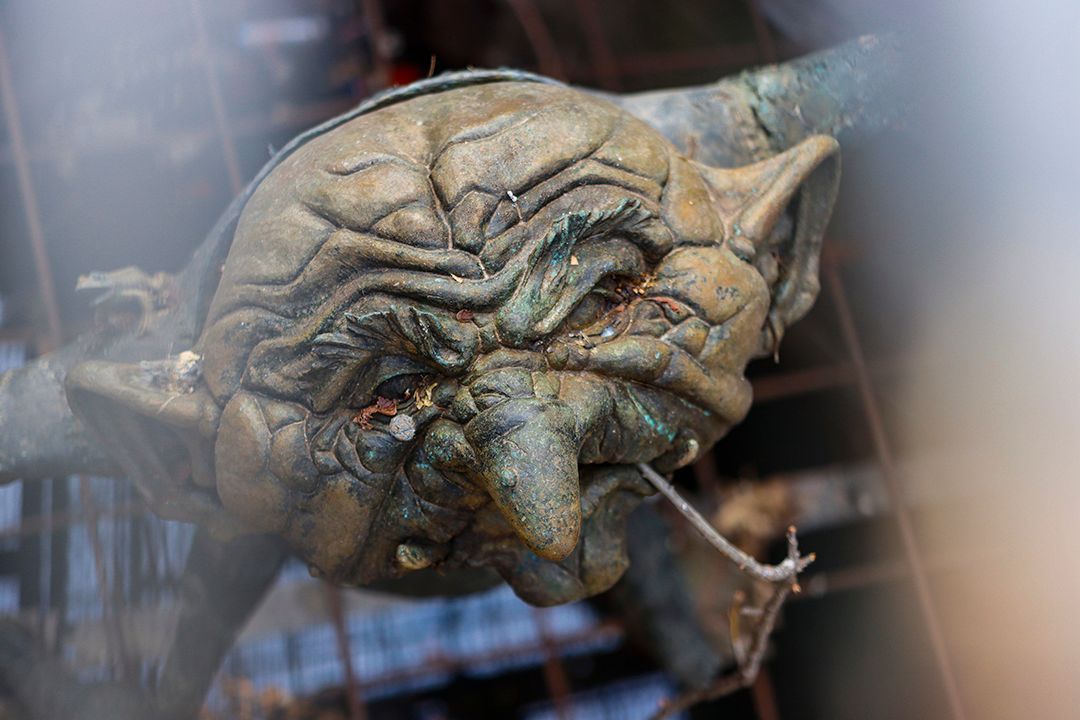A close-up of a textured, bronze-colored sculpture of a grimacing, goblin-like face with wrinkled, sagging features.