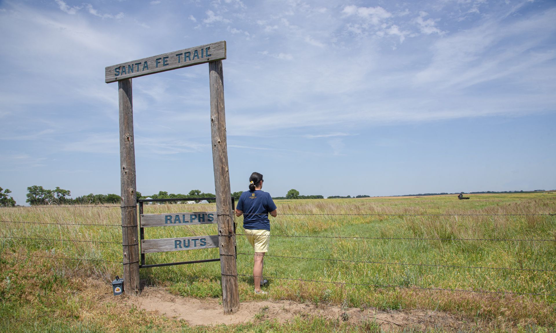 A person standing under a wooden archway in a grassy field on a sunny day.