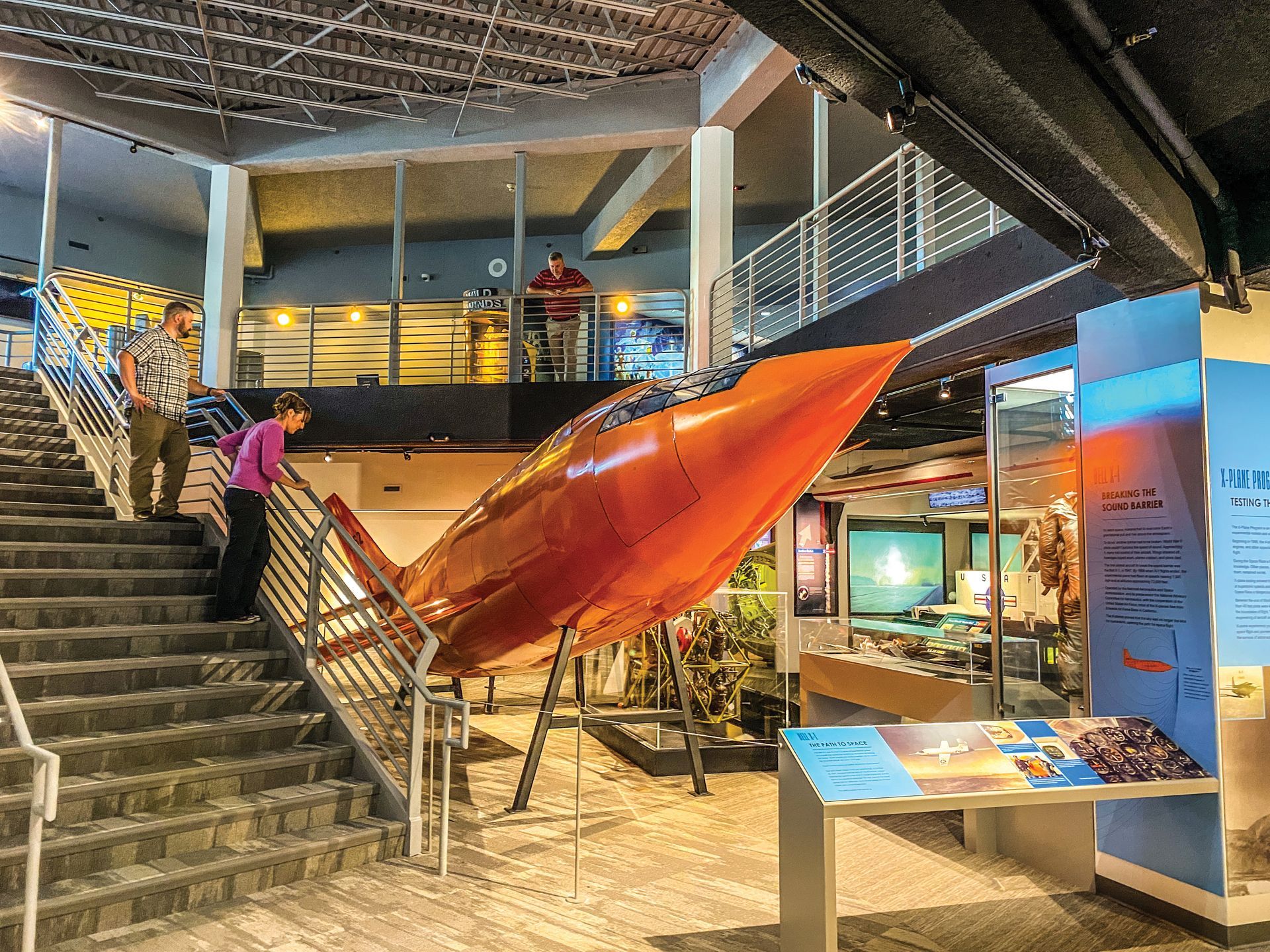 A large, orange rocket model hangs in an indoor museum exhibit next to a staircase with people observing it.