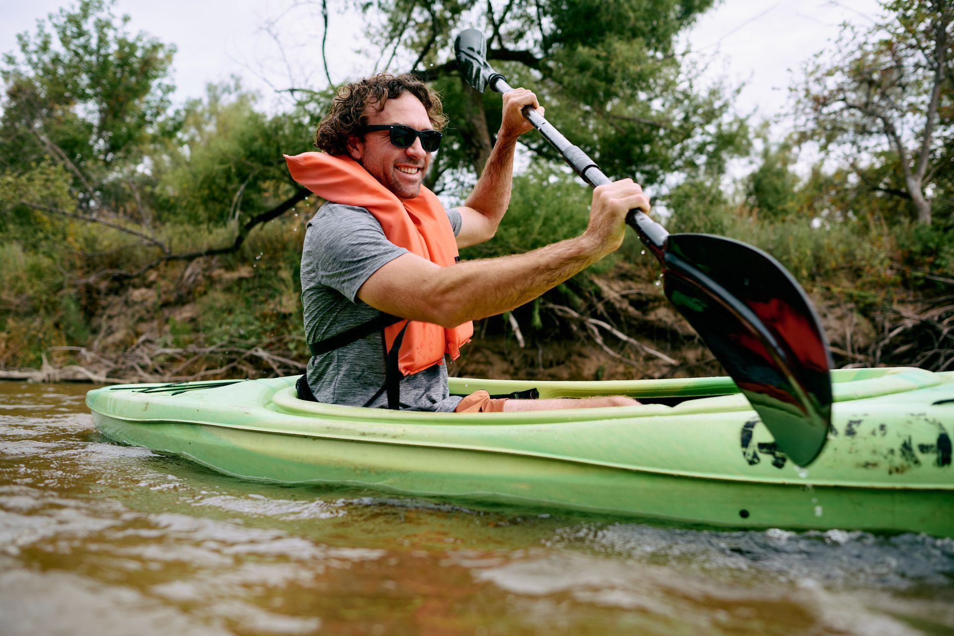 A person with curly hair and sunglasses wearing an orange life vest paddles a light green kayak on the Arkansas River.