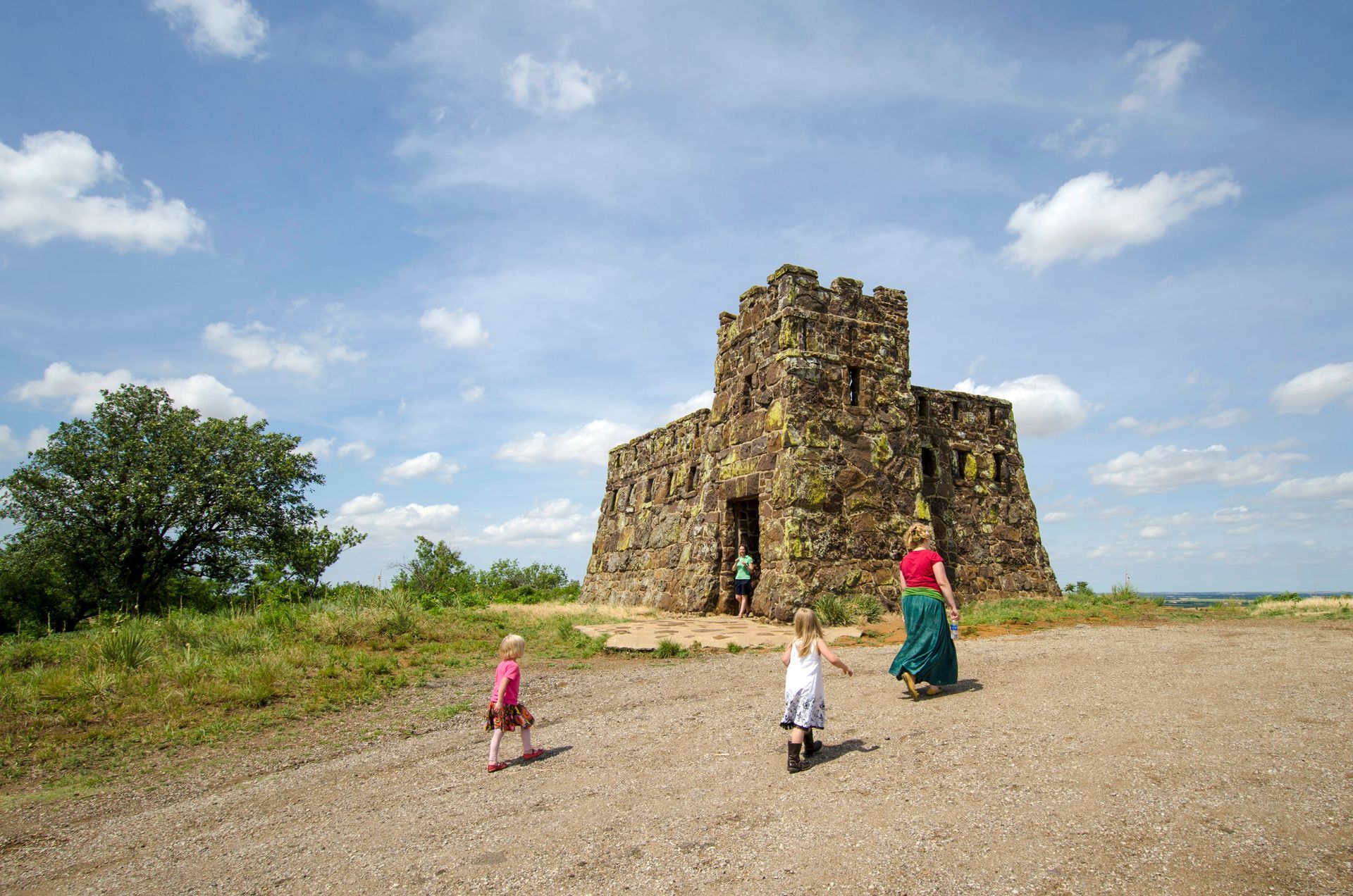 People walk toward a stone tower on a gravel path under a blue, cloudy sky.
