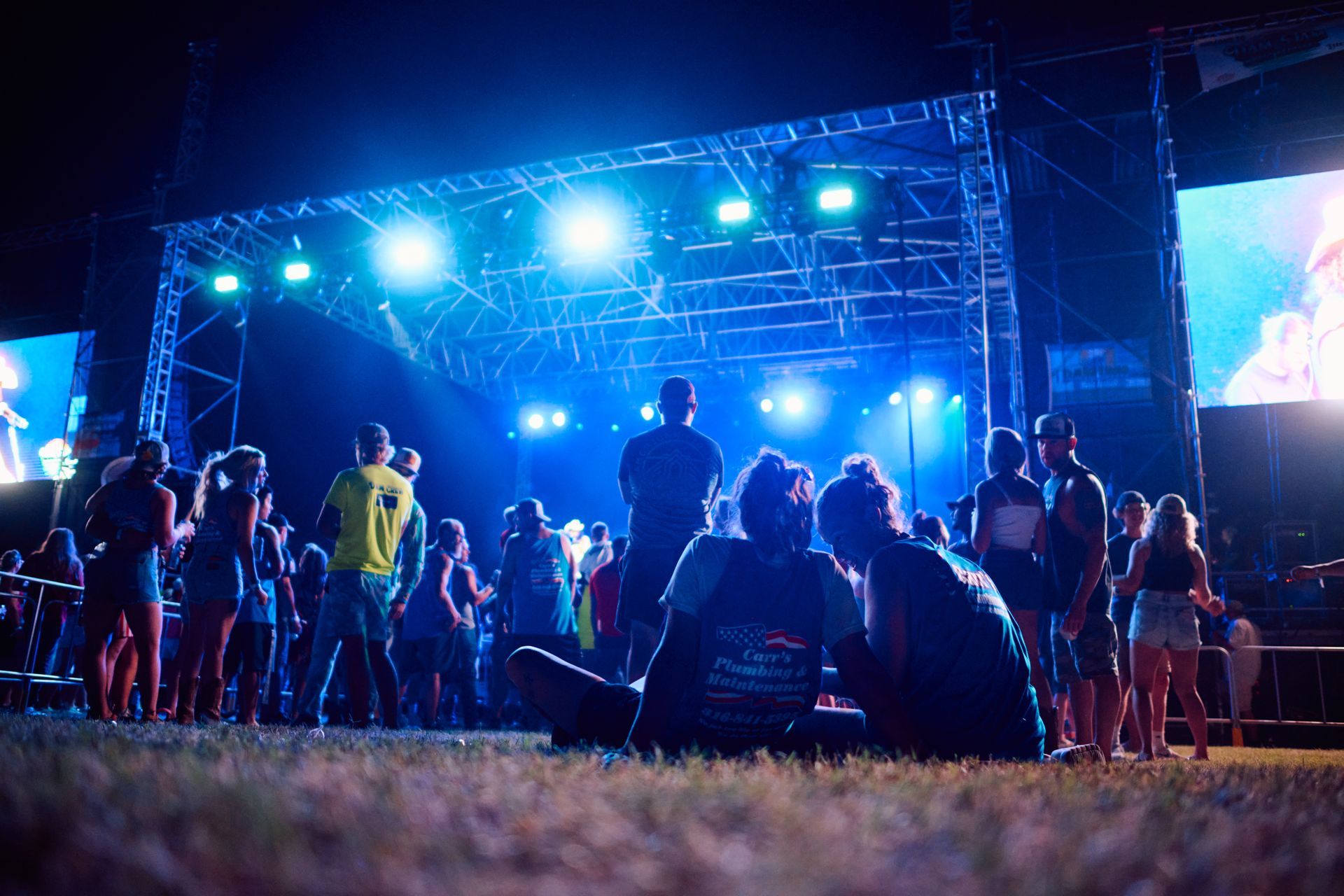 A crowd watches a concert performance at night, illuminated by bright blue stage lights and a large screen.