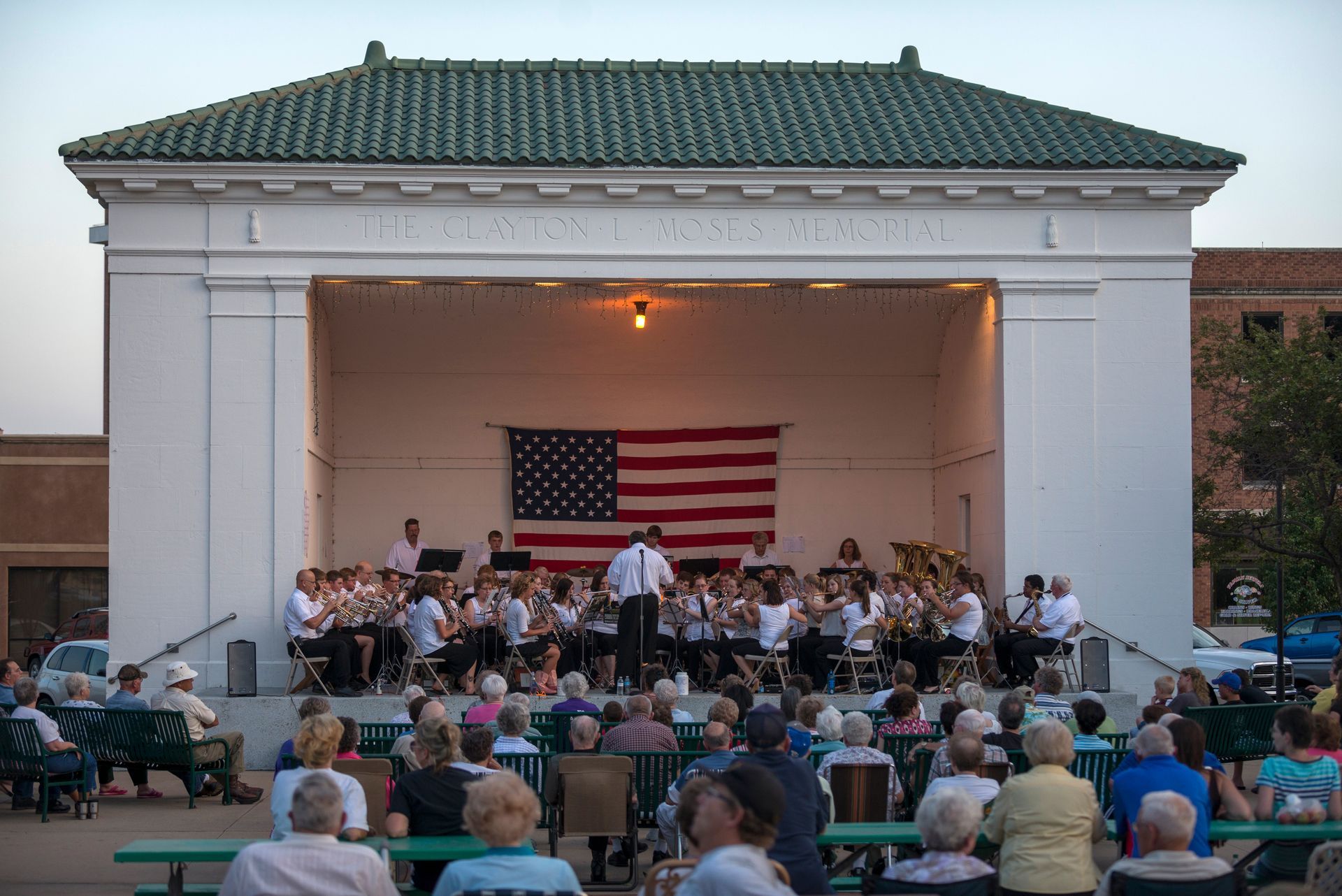 A wind ensemble performs on a stage under a gazebo with an American flag backdrop, facing an outdoor audience.
