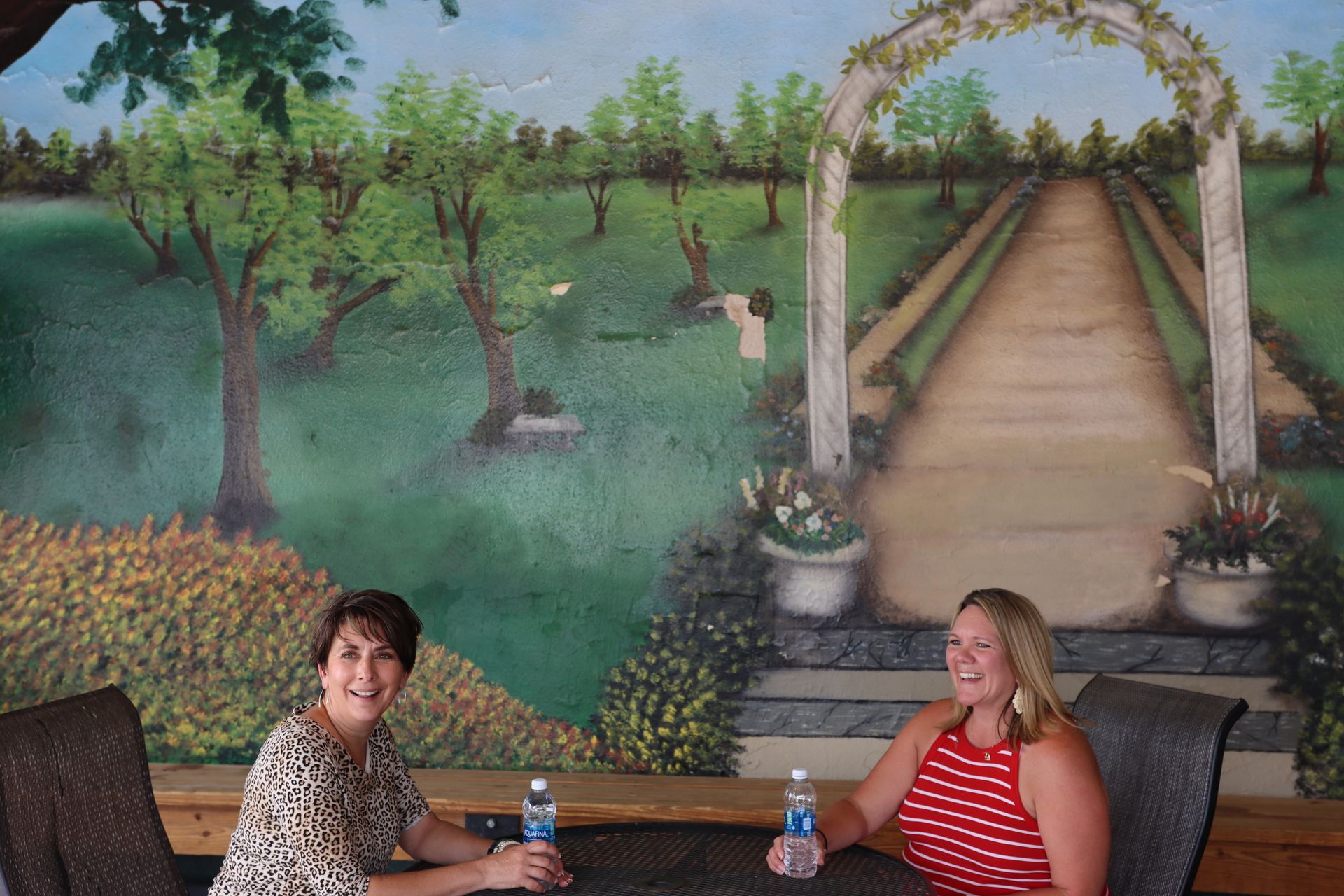 Two people sit at a table in front of a mural depicting a garden path under a trellis.