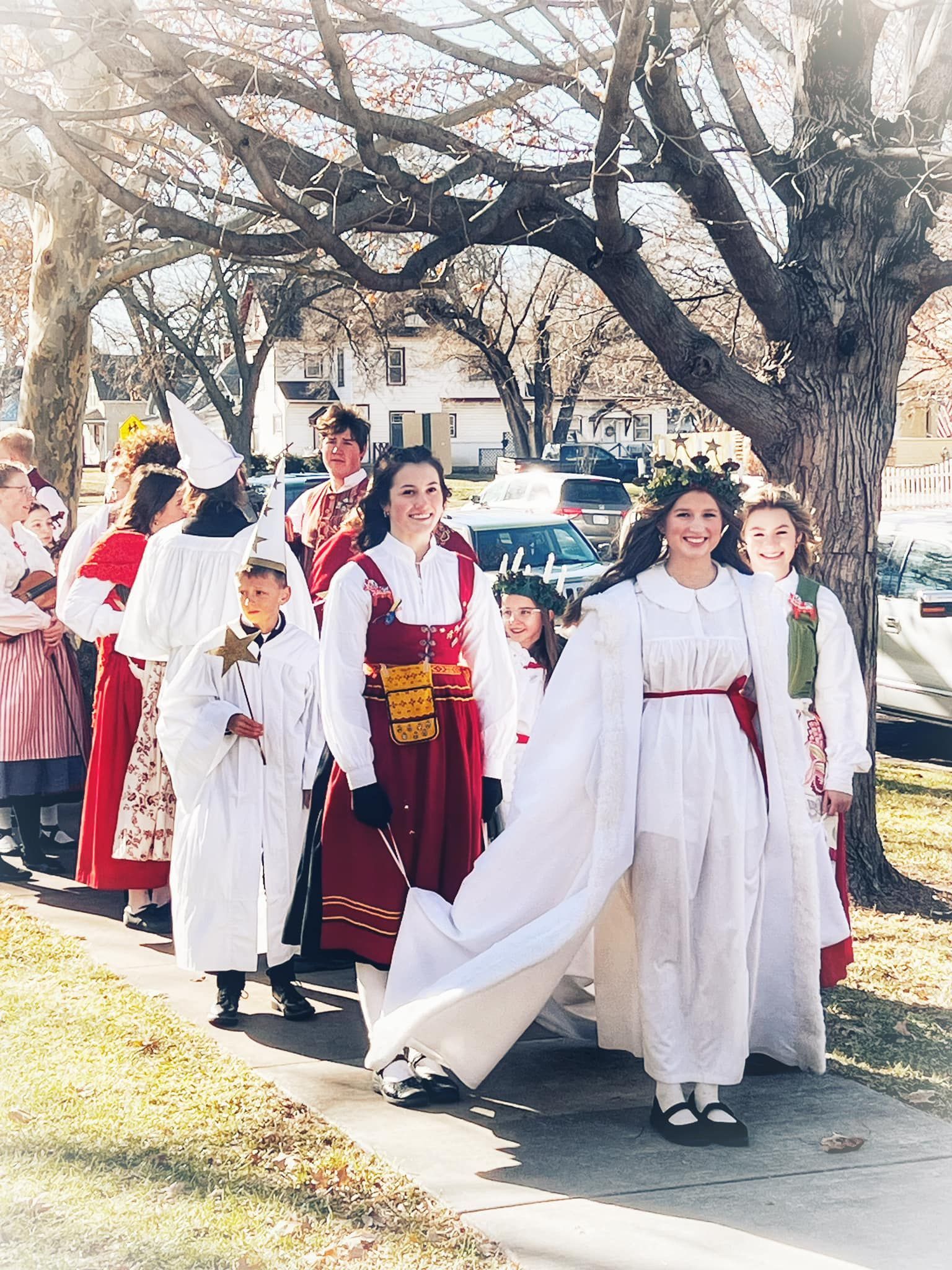 People in traditional Scandinavian Lucia procession costumes, including a candle-crowned figure, walking along a sidewalk.