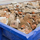 Blue metal dumpster filled with broken bricks, concrete, and rubble on a street.
